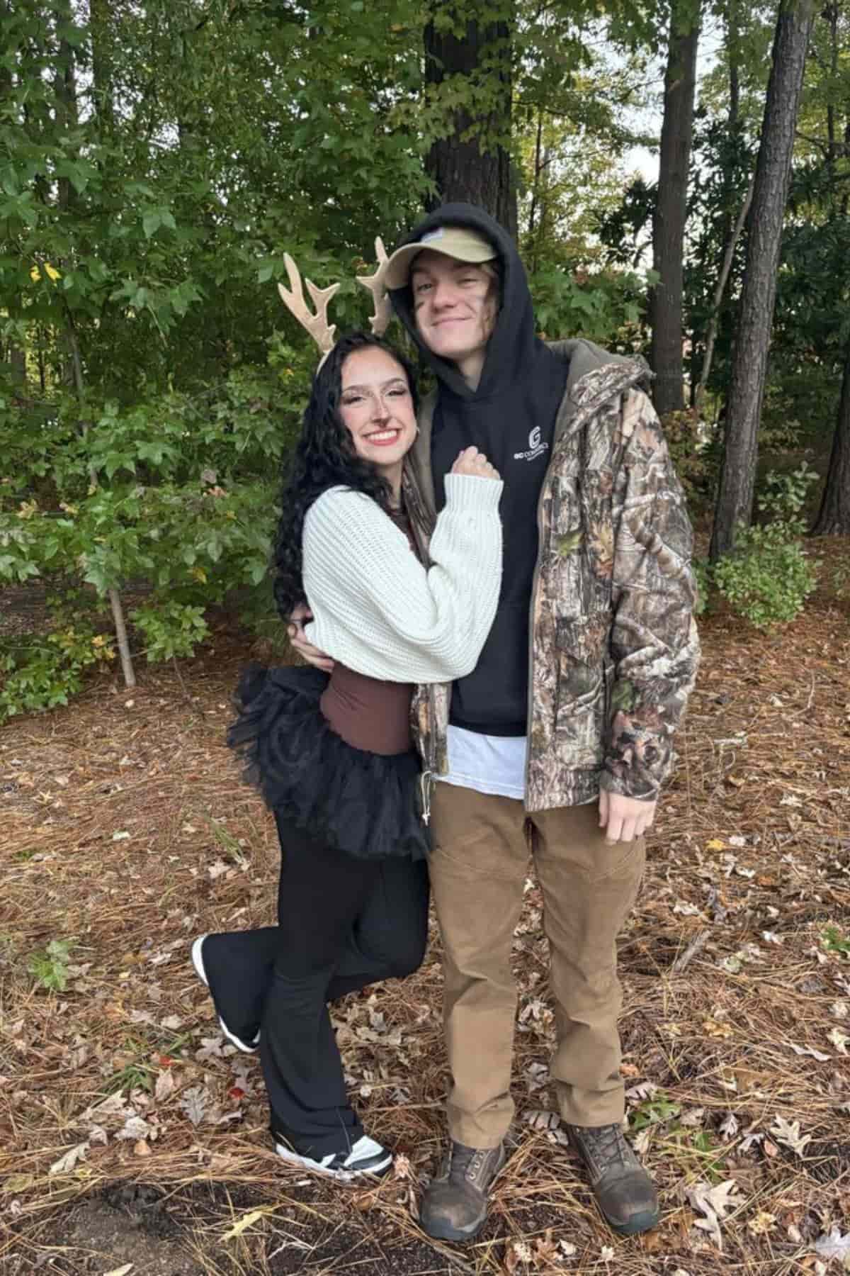 Two people pose in a wooded area; one wears antlers and a tutu, and the other wears a camouflage jacket and baseball cap.