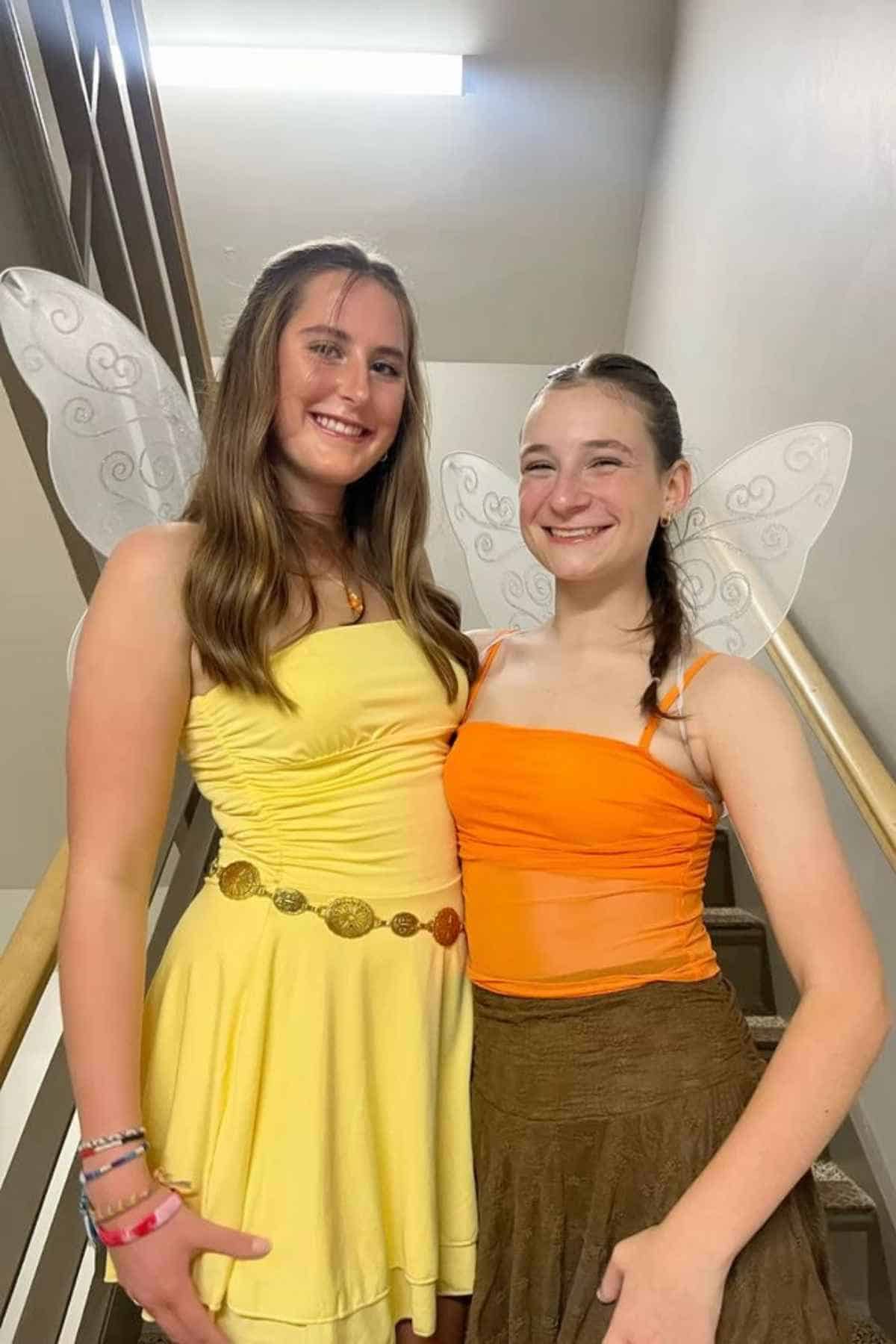 Two young women in fairy costumes with wings stand on a staircase, one in a yellow dress and the other in an orange top and brown skirt, both smiling at the camera.