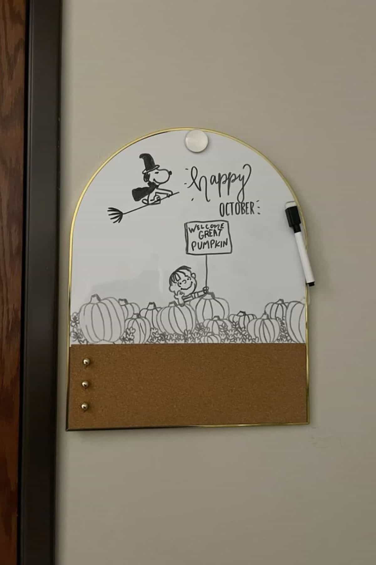 A whiteboard with Peanuts characters, Snoopy on a broomstick, “Happy October,” and “Welcome Great Pumpkin” handwritten, above a corkboard section with three pins and a marker attached.
