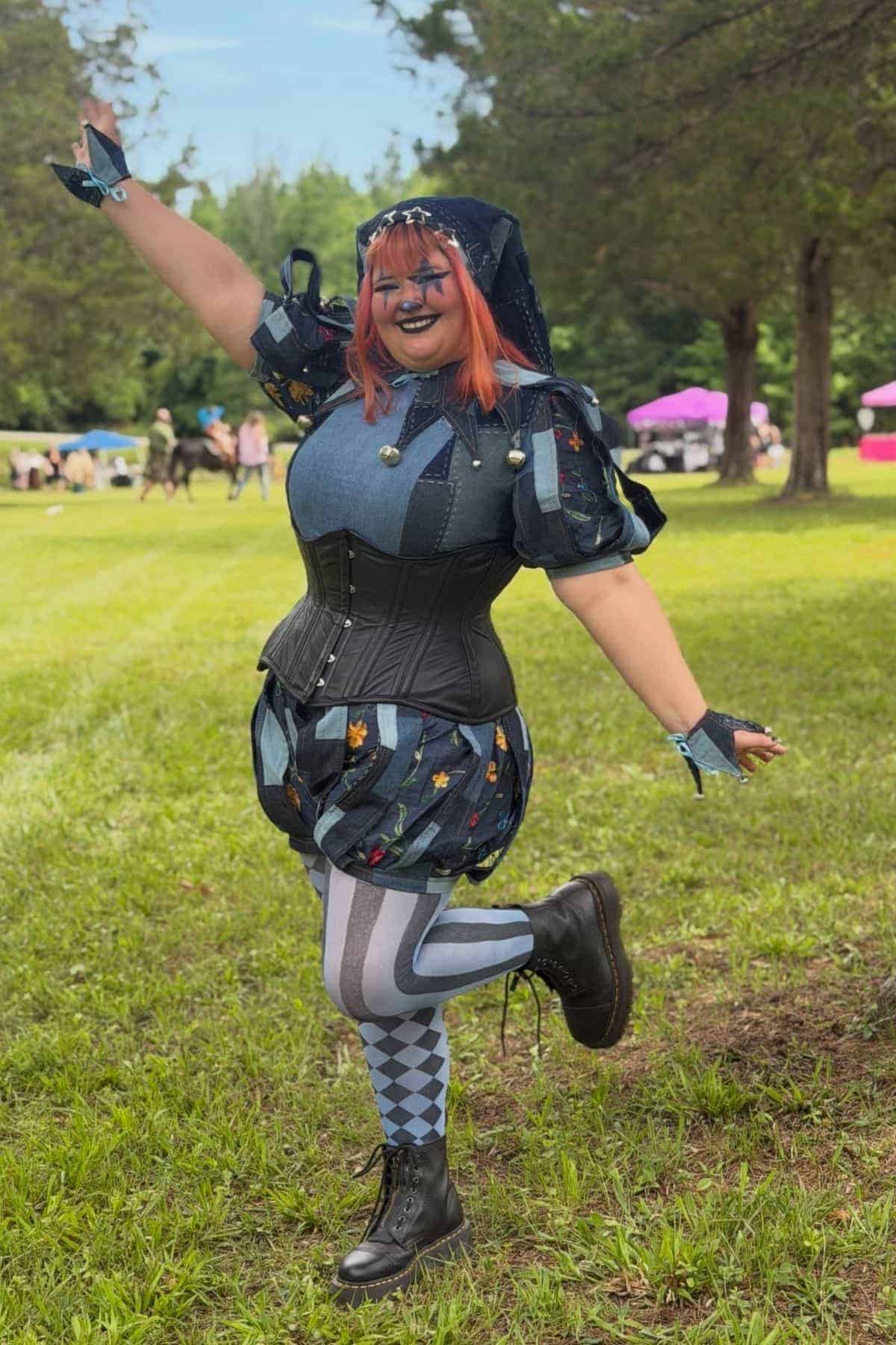 Person in a jester-inspired costume with a corset, patterned tights, and black boots poses cheerfully outdoors on grass with trees and people in the background.