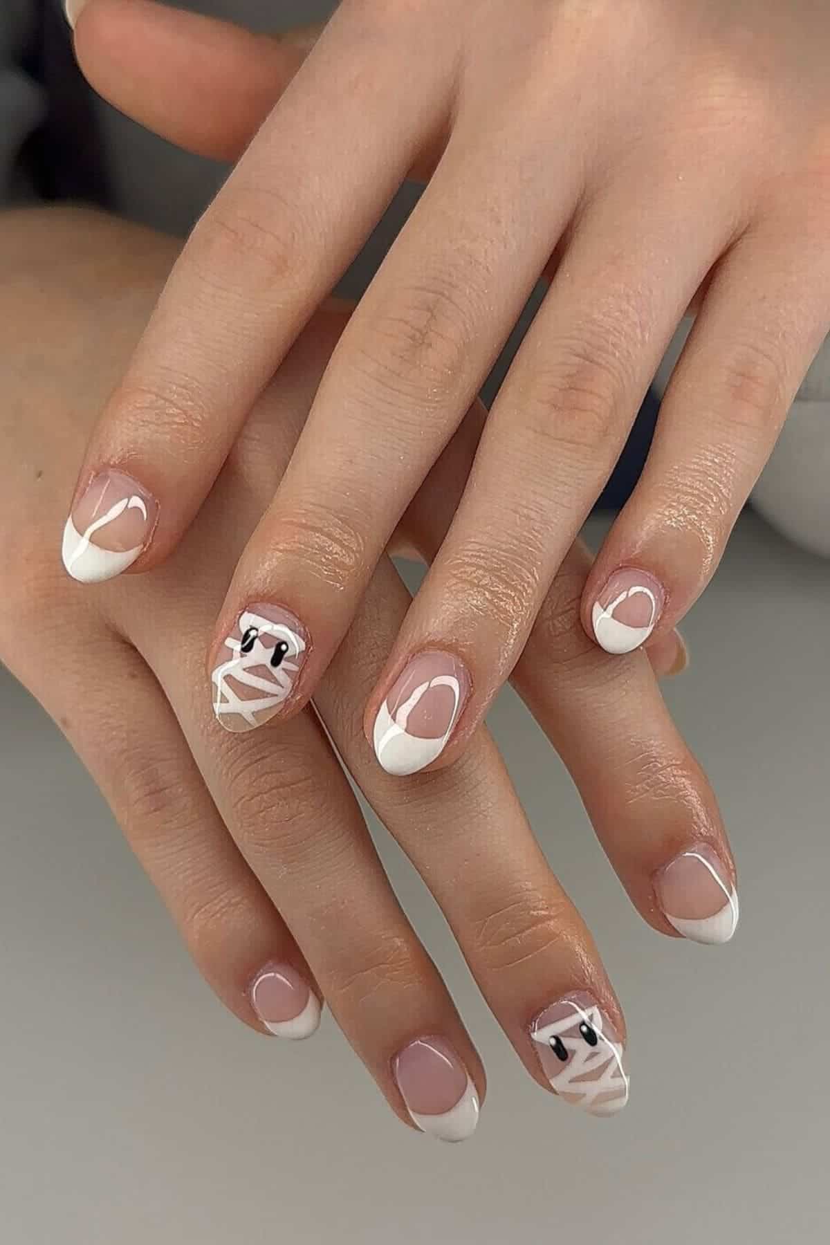 Hands with French manicured nails; two nails feature white mummy designs with black and white eyes, suitable for a Halloween theme.