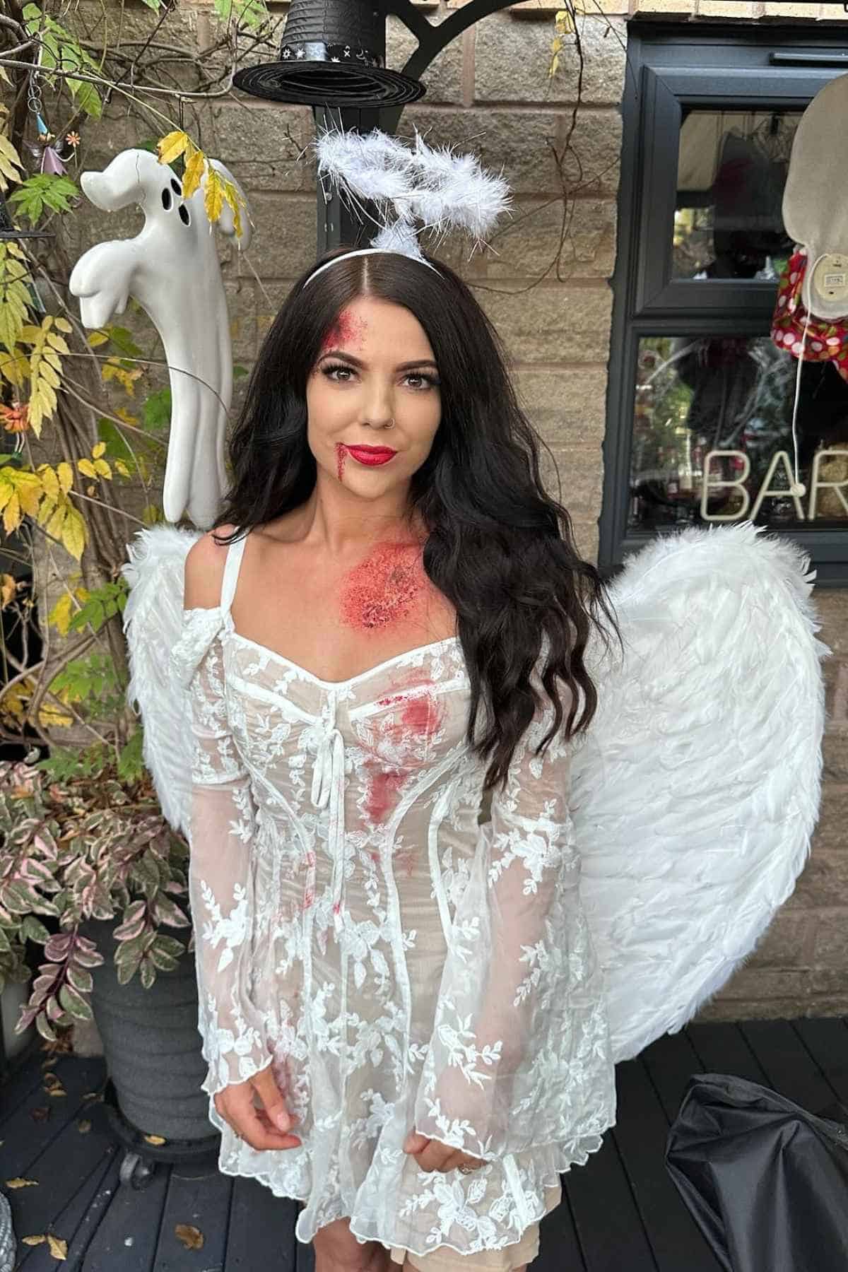 Woman wearing a white lace angel costume with wings, halo, fake blood, and makeup wounds, standing outdoors near plants and Halloween decorations.