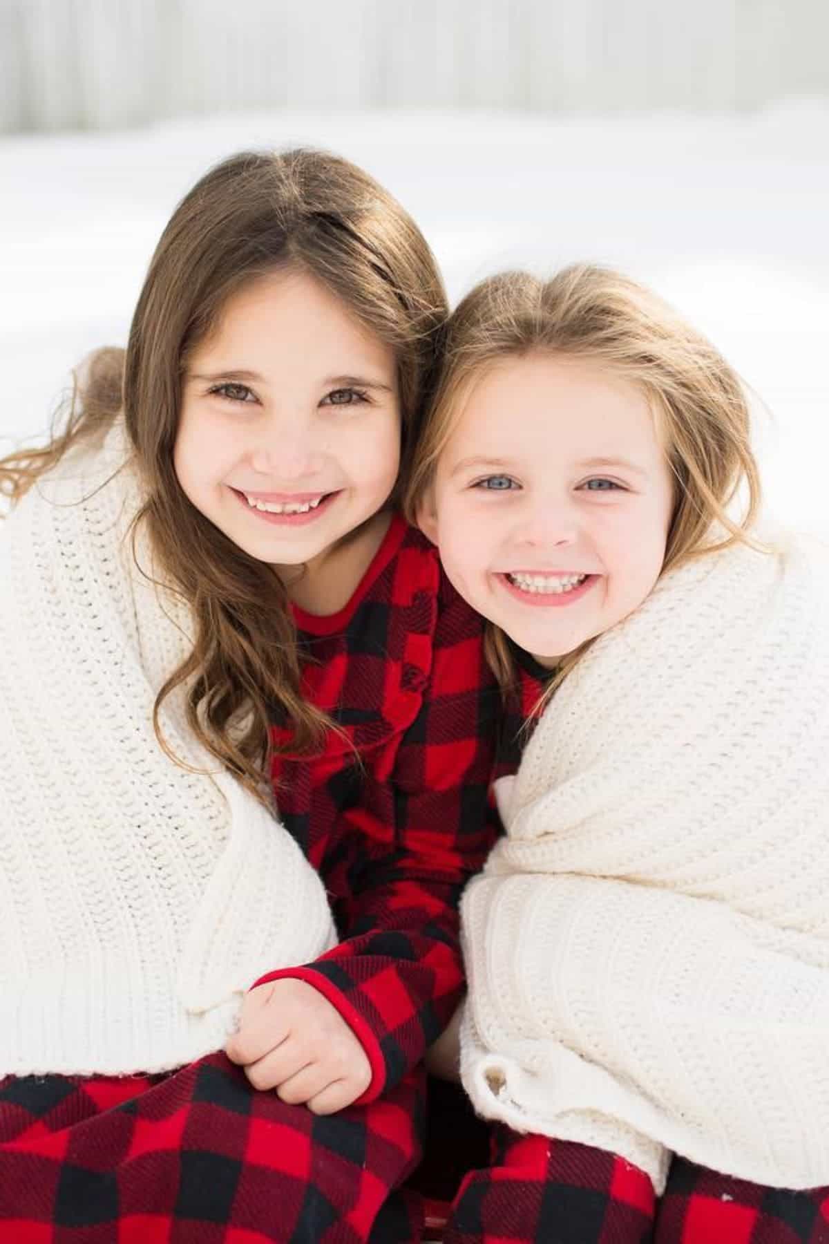 Two young girls in red and black plaid outfits sit together outside, smiling at the camera and sharing white knit blankets, with snow visible in the background.