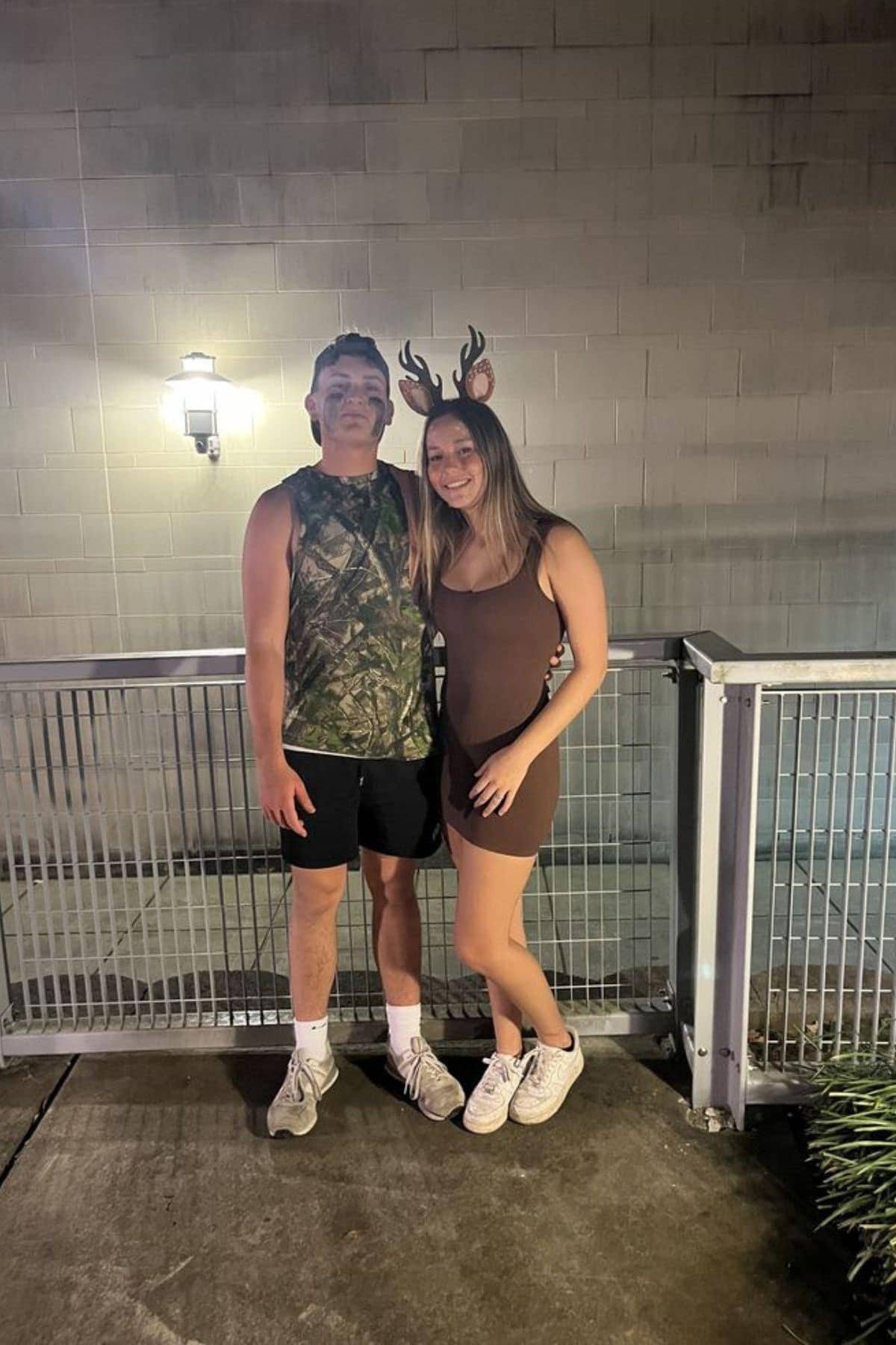 Two people stand side by side in costumes; one wears camouflage with face paint, the other wears brown with antlers and deer ears, posing outdoors by a metal railing at night.