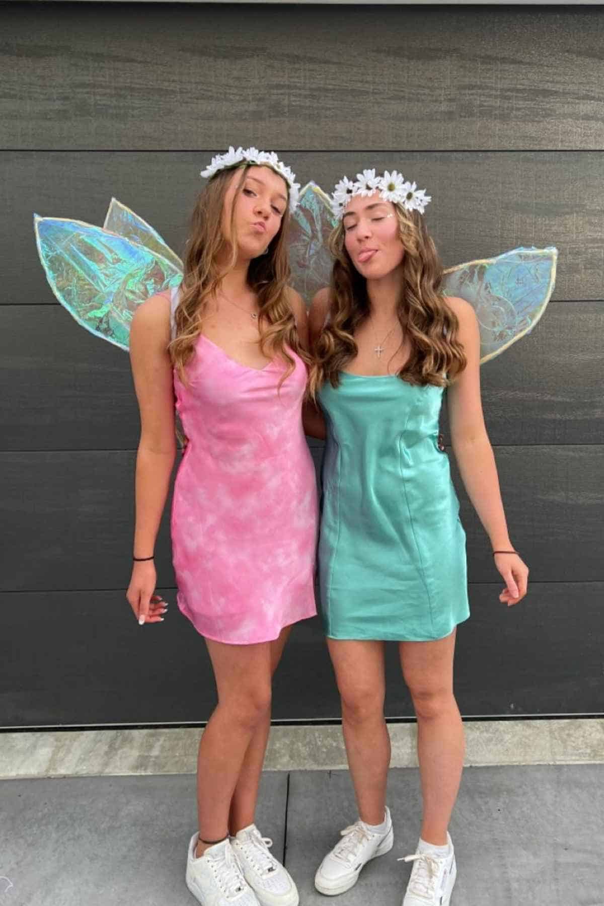 Two young women wearing pastel dresses, fairy wings, white sneakers, and flower crowns stand side by side, facing forward and sticking out their tongues.