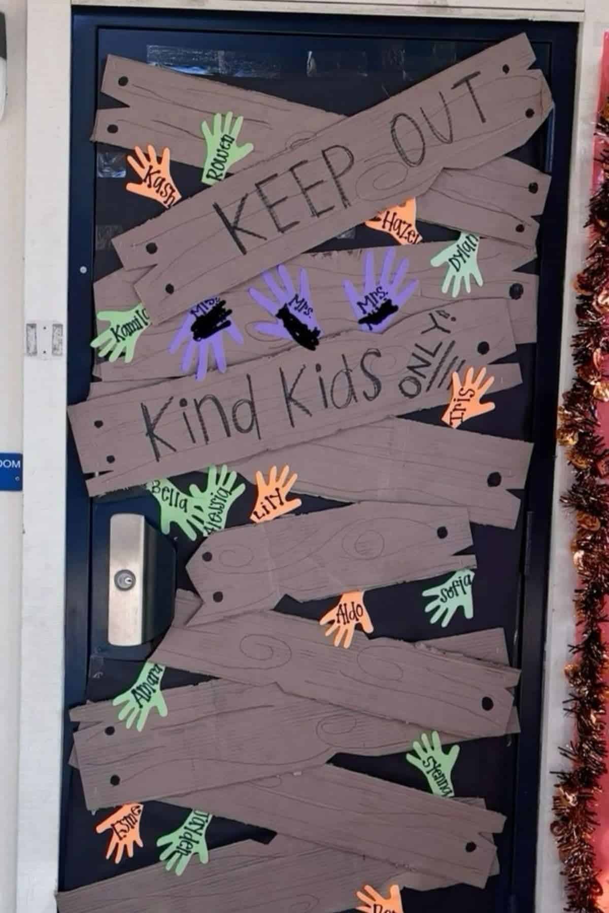 A classroom door decorated with paper boards and handprints, reads “KEEP OUT” and “Kind Kids ONLY,” with children’s names written on colorful paper hands.