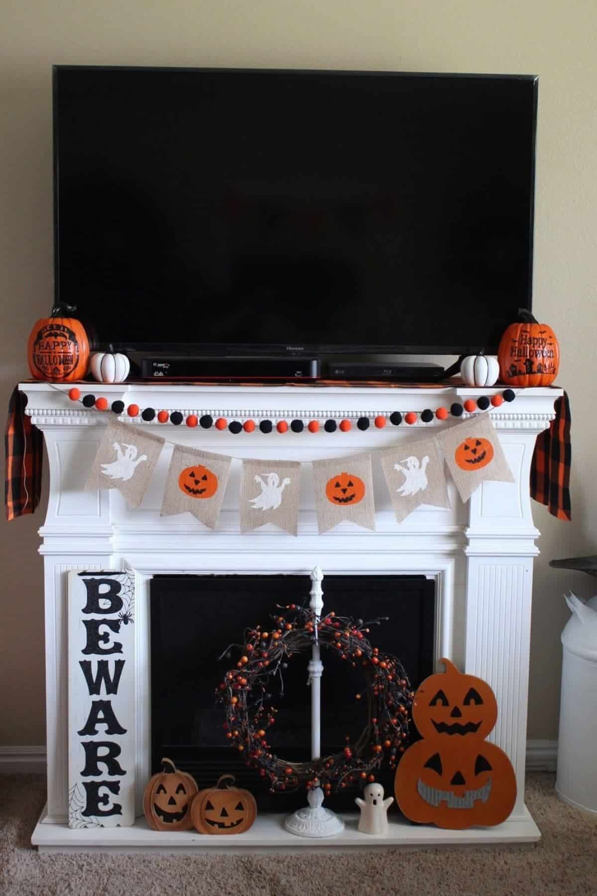 A white mantel decorated with Halloween items, including pumpkins, garlands, and banners, sits below a TV. A “BEWARE” sign and a wreath with orange lights complete the setup.