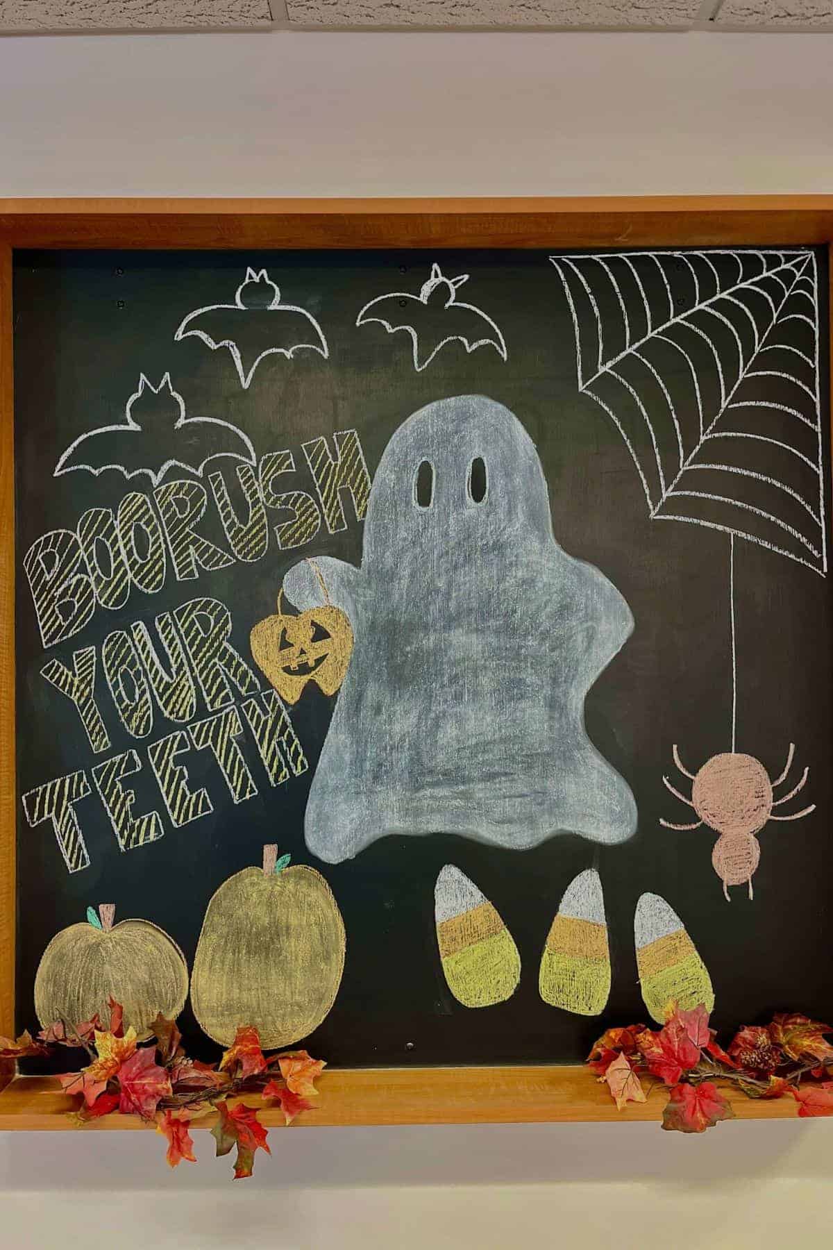 A chalkboard drawing of a ghost holding a pumpkin with “BOOrush YOUR TEETH” text, bats, candy corn, spider web, spider, pumpkins, and autumn leaves.