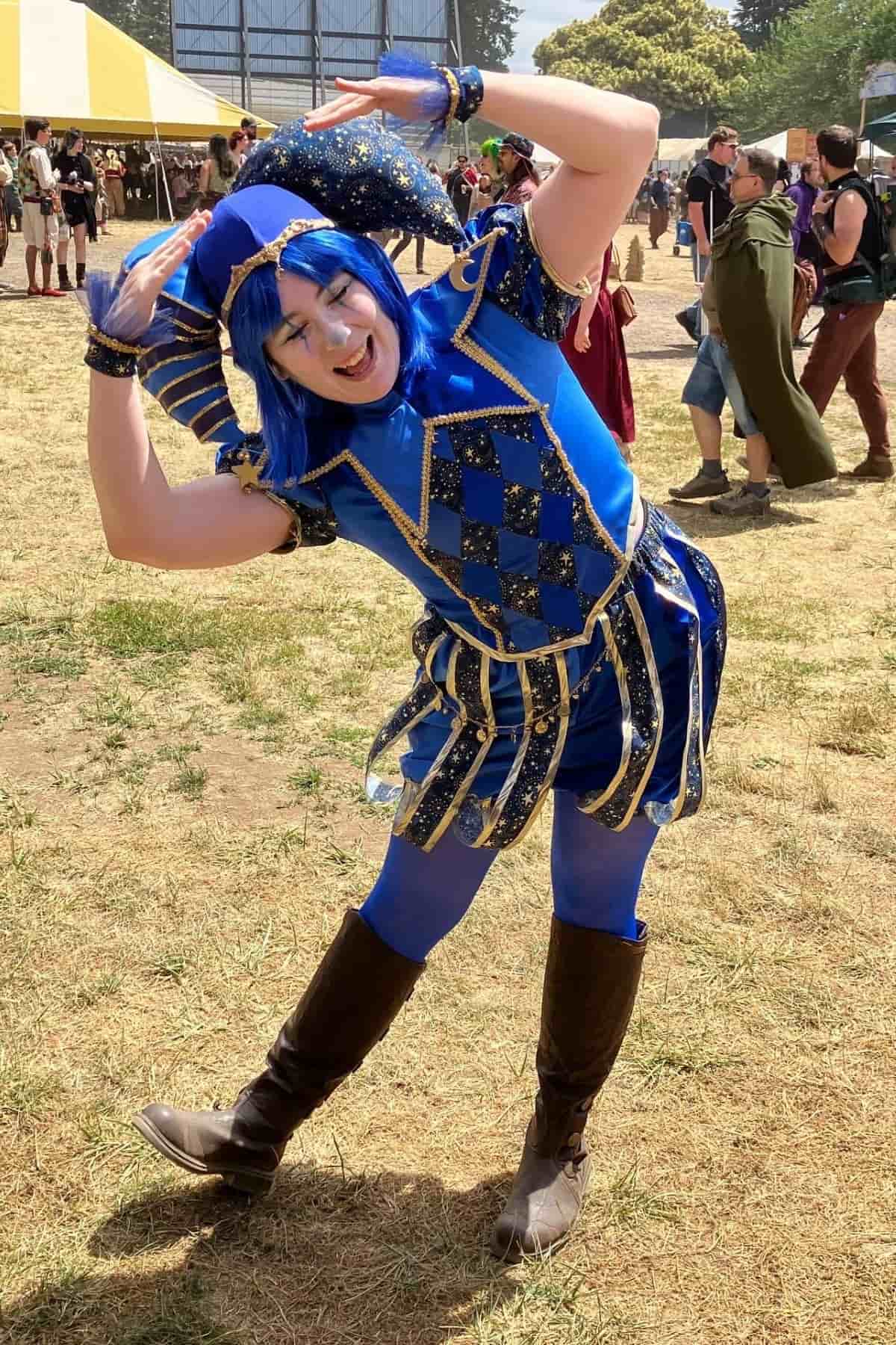 Person in a blue jester costume with gold trim and boots poses outdoors at a festival, tilting to one side with arms raised, surrounded by other attendees.