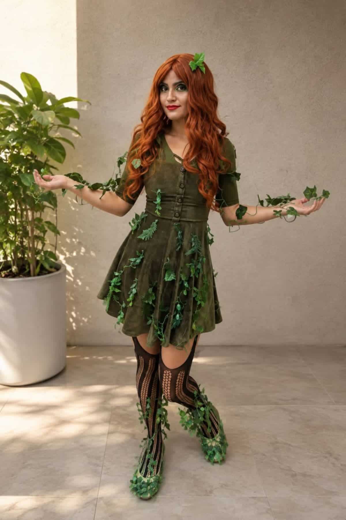 Woman in a green dress with red hair, decorated with ivy and leaves, stands indoors near a potted plant, posing with arms slightly raised.