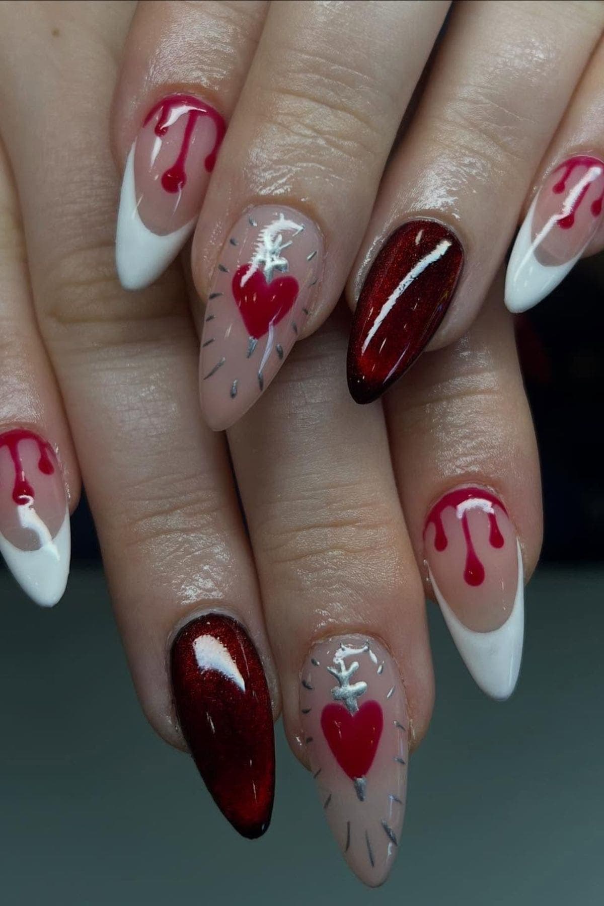 Hands with long, pointed nails featuring designs of red hearts, white stitching, dripping blood patterns, and solid red and white accents.