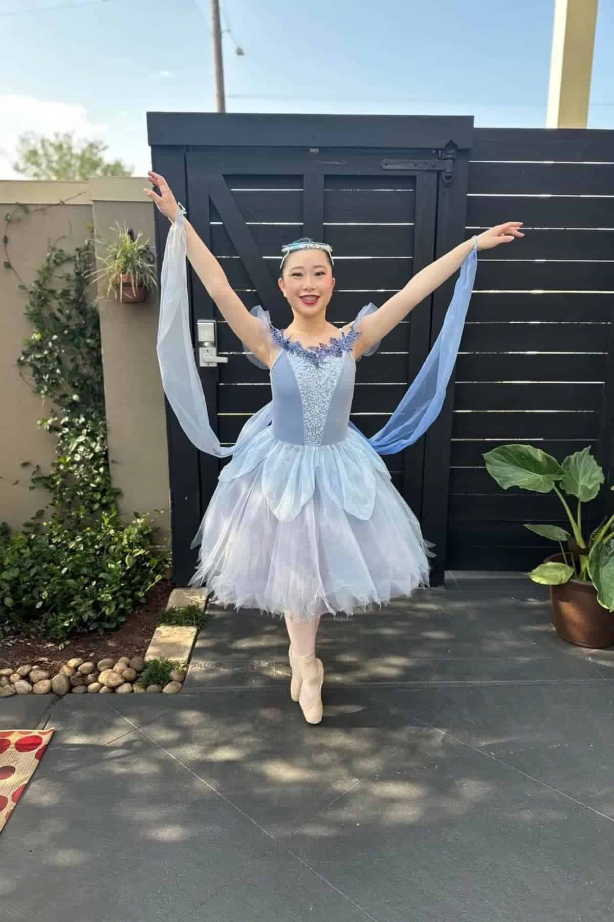 A ballerina in a blue tutu poses en pointe with arms raised outdoors on a patio near plants and a black gate.