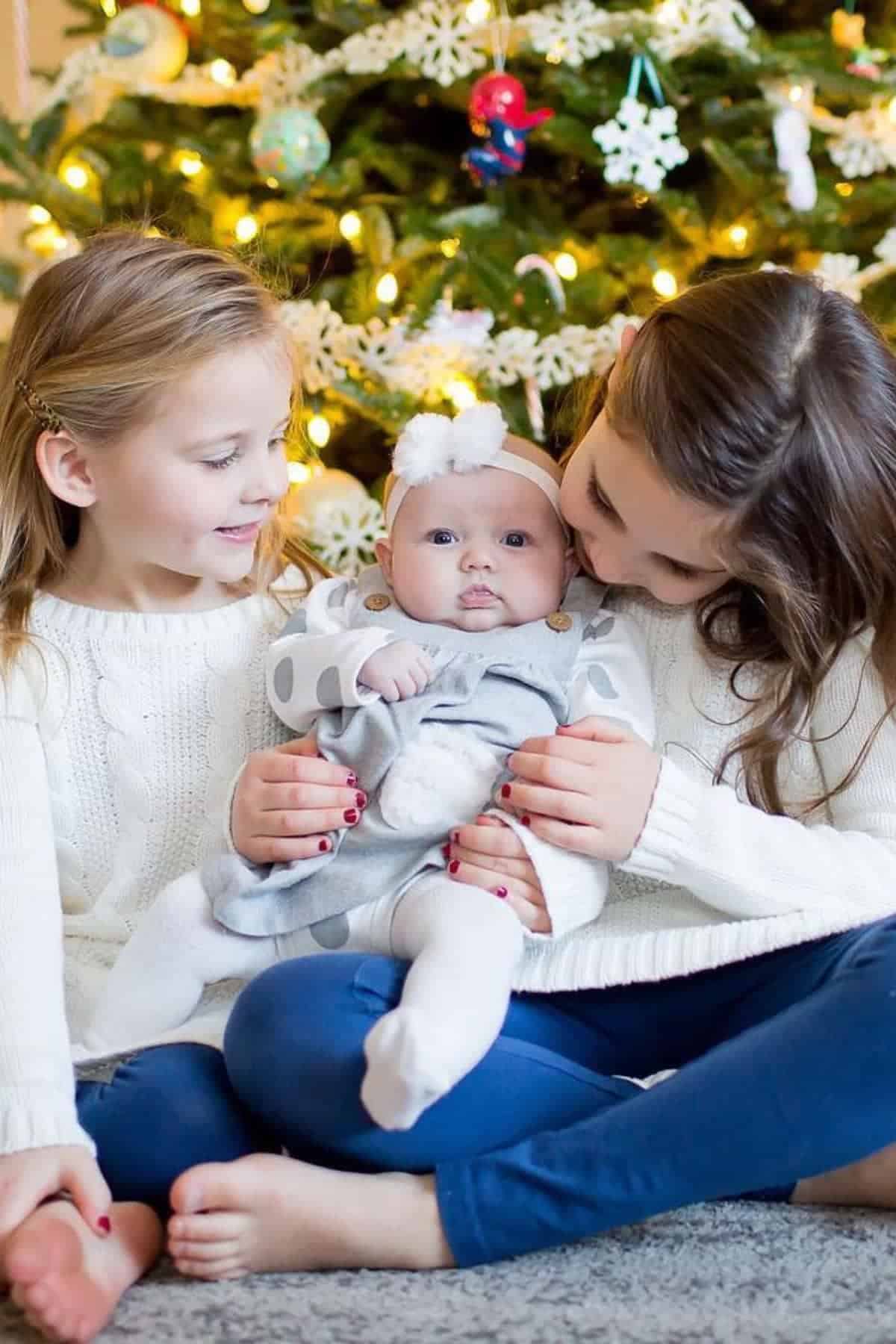 Two young girls sit in front of a decorated Christmas tree, holding a baby dressed in gray and white, all wearing cozy sweaters and smiling.