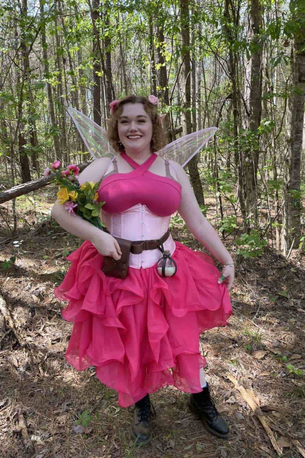 Person in a pink fairy costume with wings, holding a bouquet of flowers, standing in a forest with sunlight filtering through the trees.
