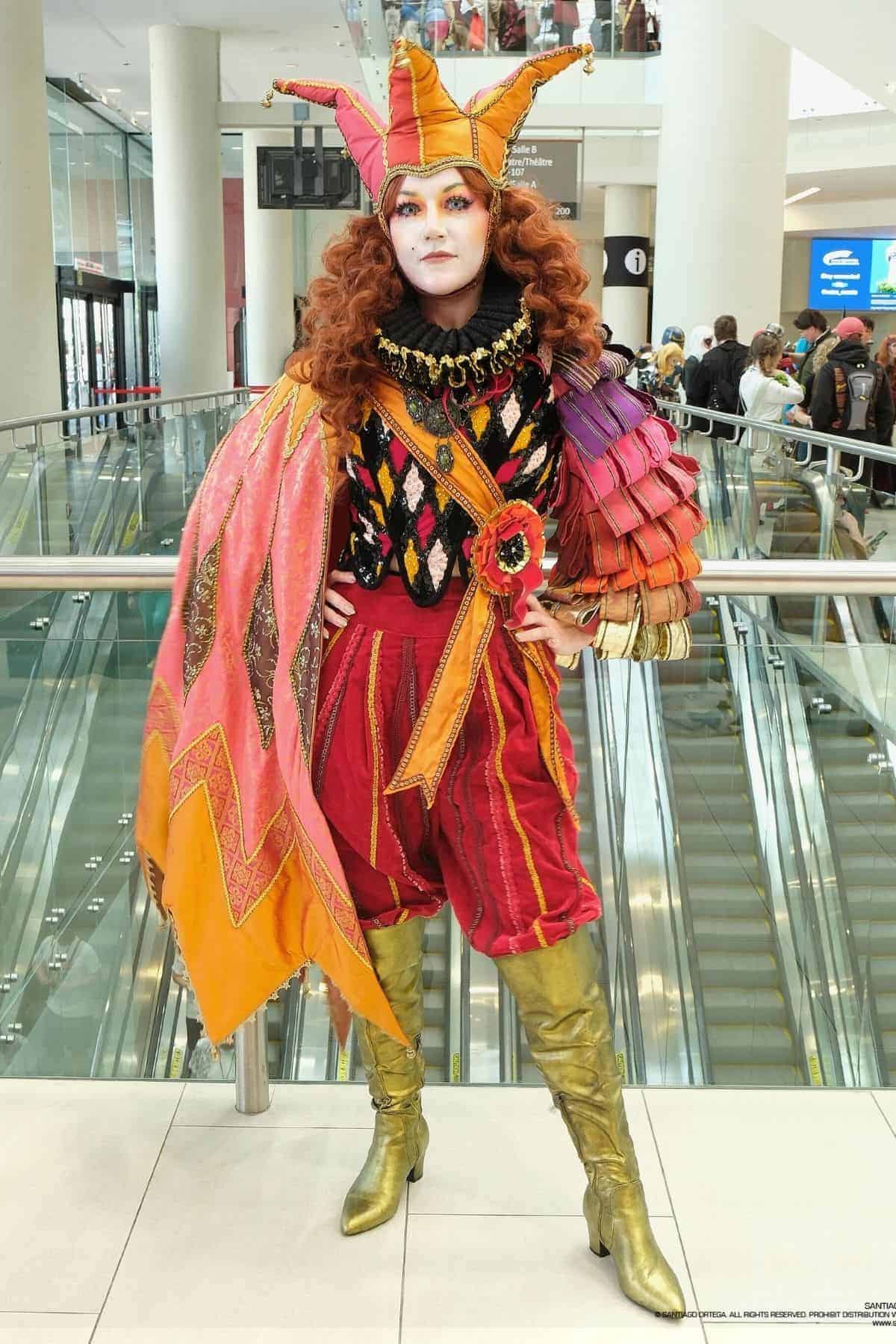 Person in a colorful jester-inspired costume with gold boots, red shorts, a multicolored tunic, ruffled collar, and a jester hat, standing indoors near escalators.