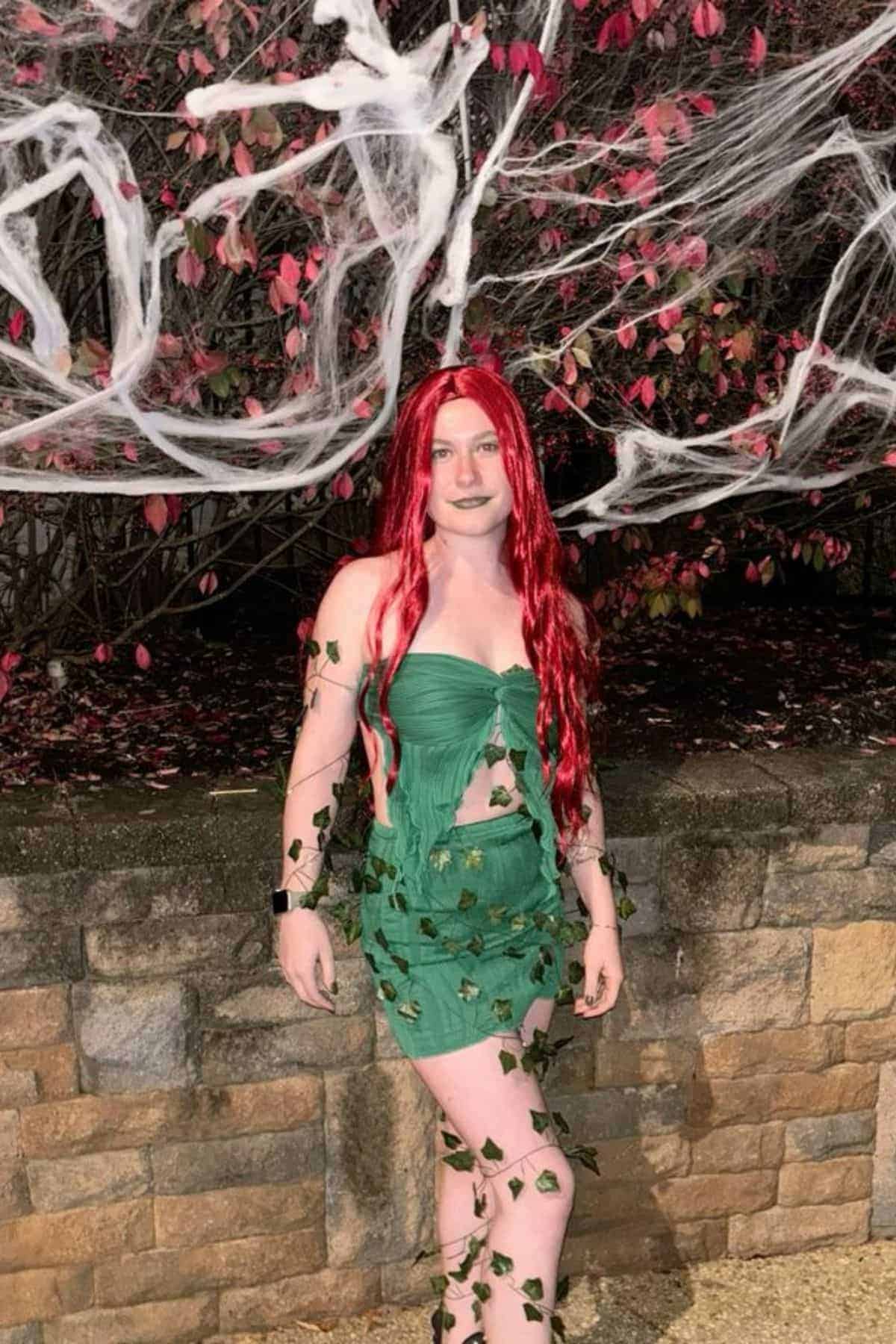 Person wearing a green leafy costume and long red wig stands in front of a stone wall with fake cobwebs and red-leaved bushes in the background.