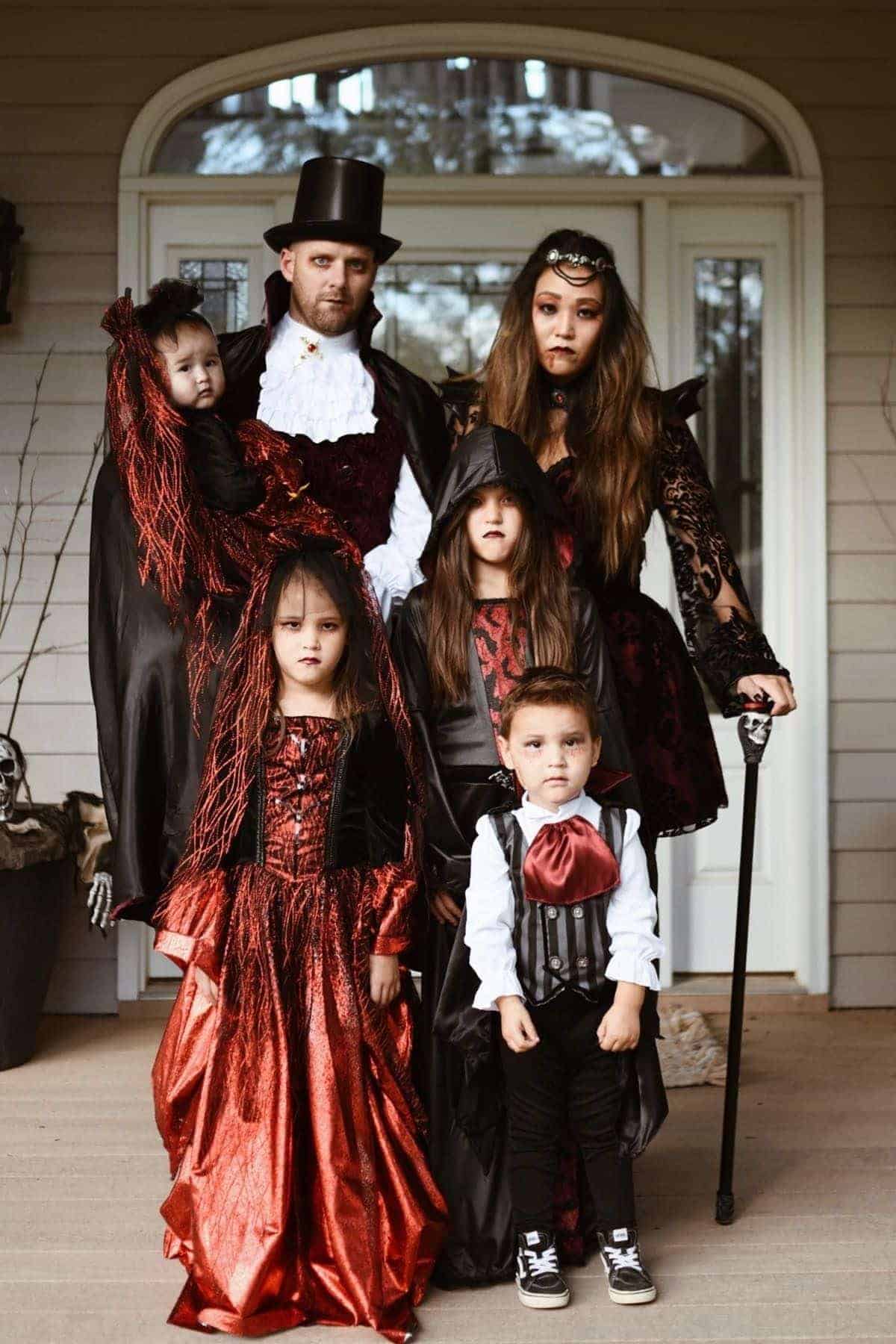 Family of six in matching gothic vampire costumes posing on a porch, with adults and children wearing black and red outfits, capes, and dramatic makeup.
