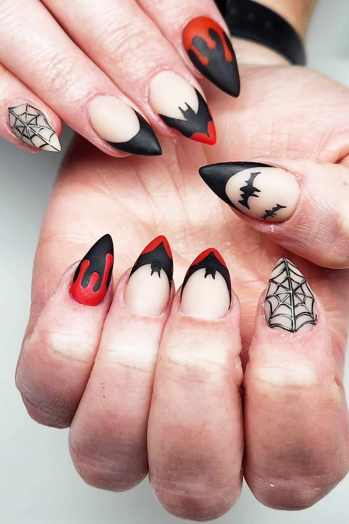 Hands with pointed nails featuring Halloween-themed nail art, including spider webs, bats, black tips, and red dripping designs on a matte nude base.