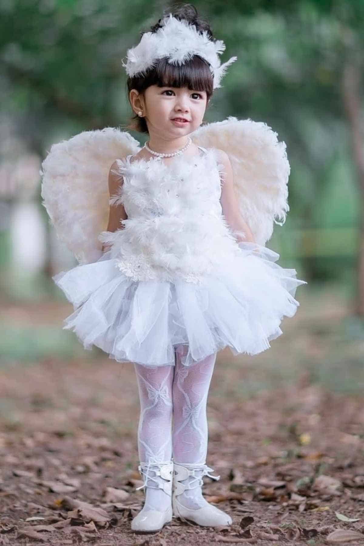 Young child in a white angel costume with wings, headband, pearl necklace, and tutu dress, standing outdoors on a leafy ground.