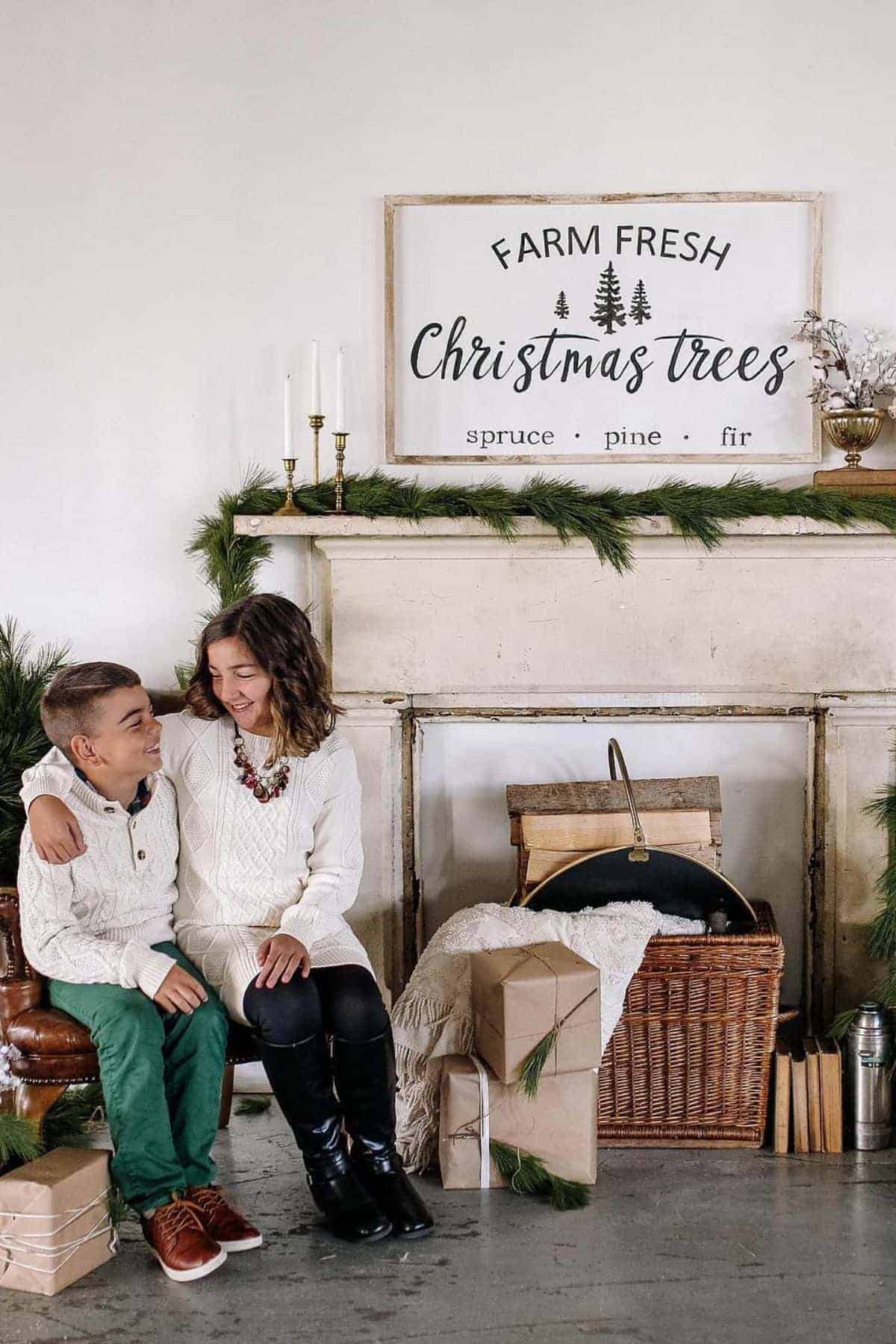 Two children sit together by a decorated fireplace with wrapped gifts, a wicker basket, greenery, and a sign that reads “Farm Fresh Christmas Trees: spruce, pine, fir.“.