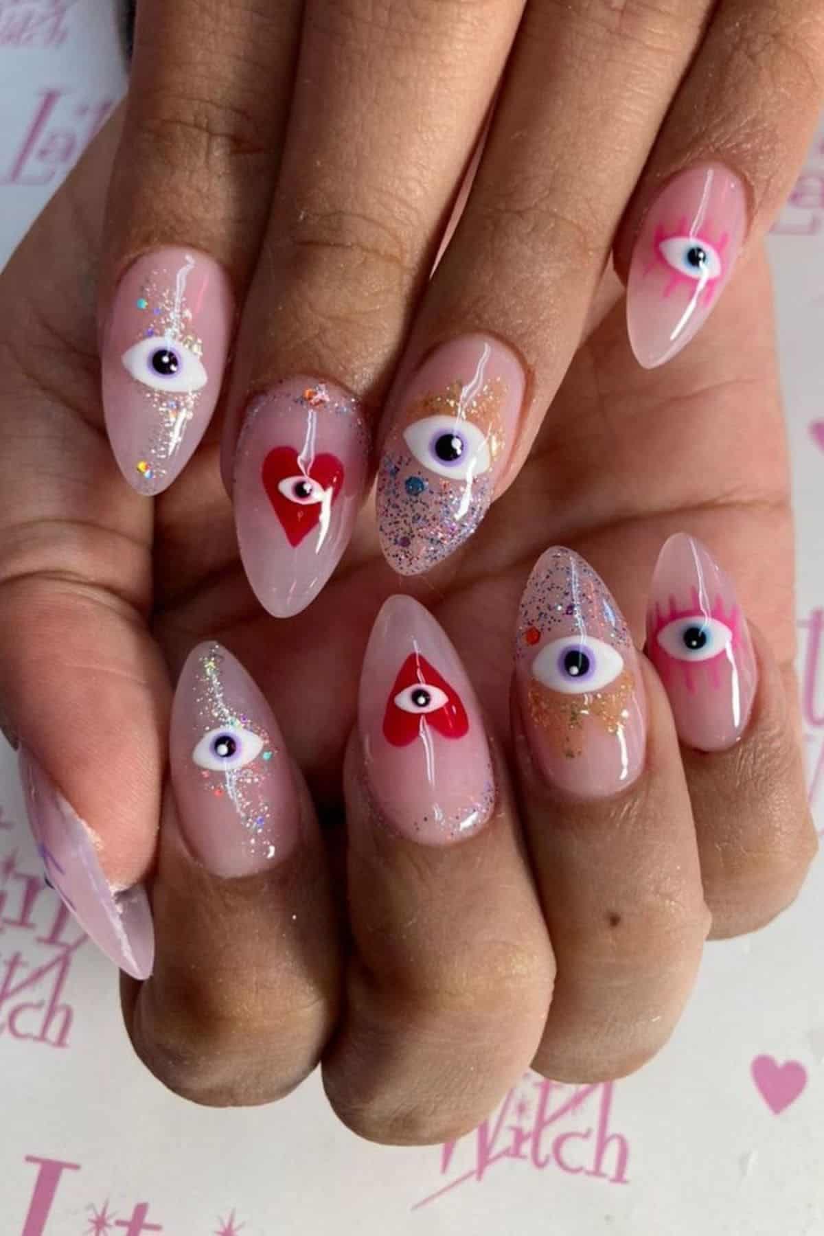 Close-up of hands with long, almond-shaped nails featuring eye and heart designs, pink and blue accents, and glitter on a nude base.