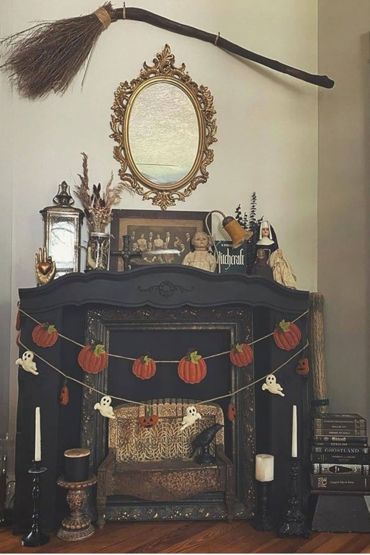 A black mantel decorated with pumpkin and ghost garlands, candles, books, a broom above a mirror, and witch-themed decor in a cozy room.