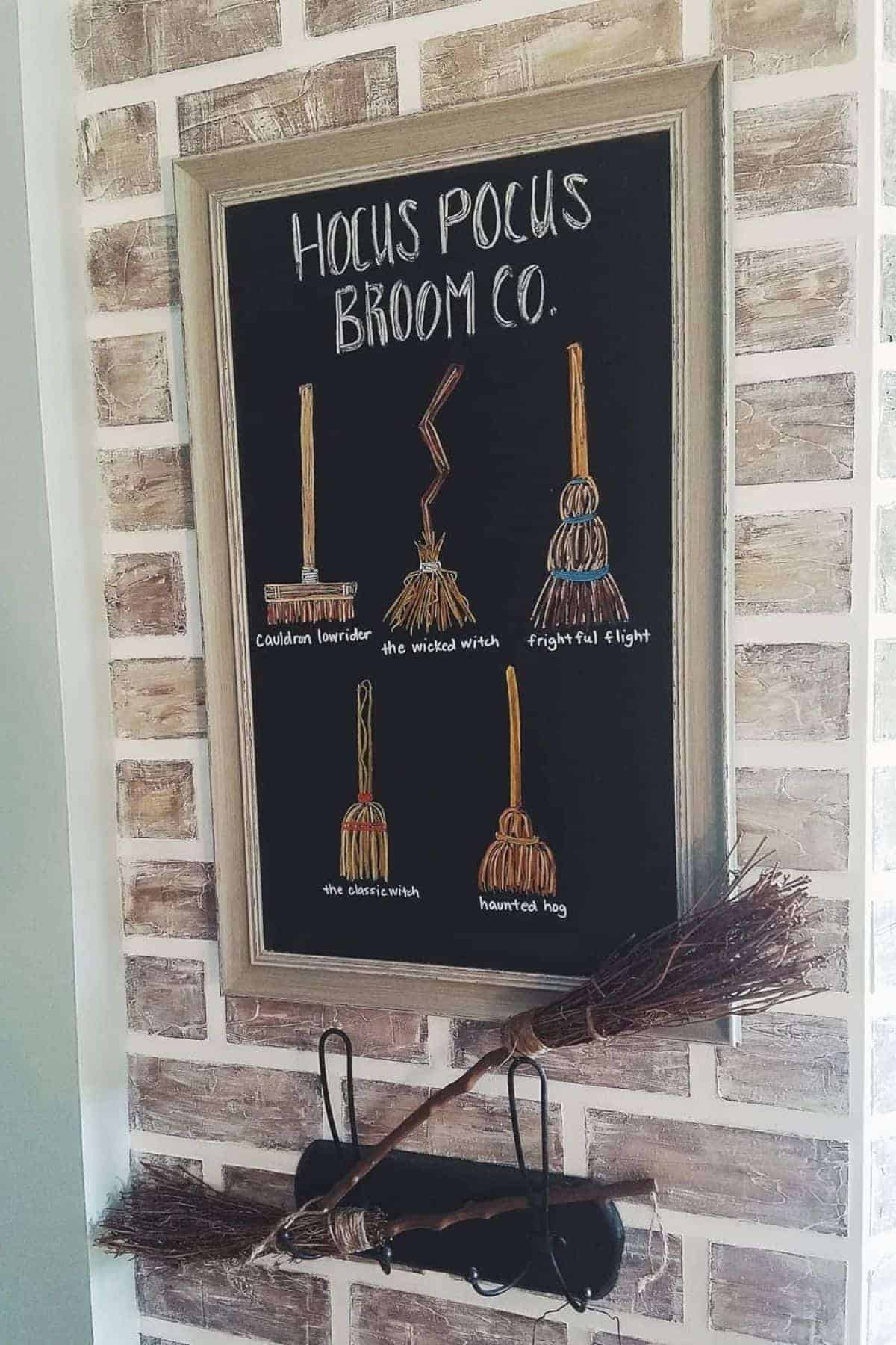 Framed sign reading “Hocus Pocus Broom Co.” with illustrated brooms and names, above two brooms hanging on a wall with a brick pattern.