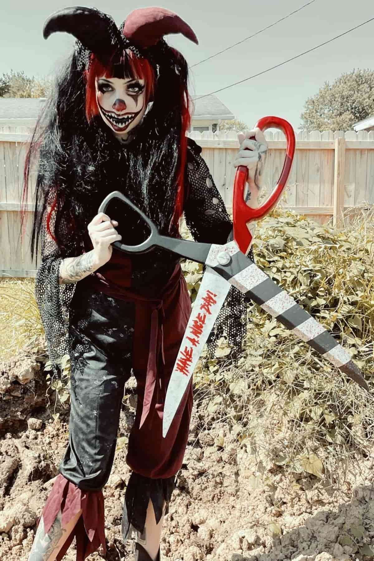 Person in black and red clown costume with eerie face makeup stands outdoors holding a large prop scissor with red handles and fake blood on the blades.