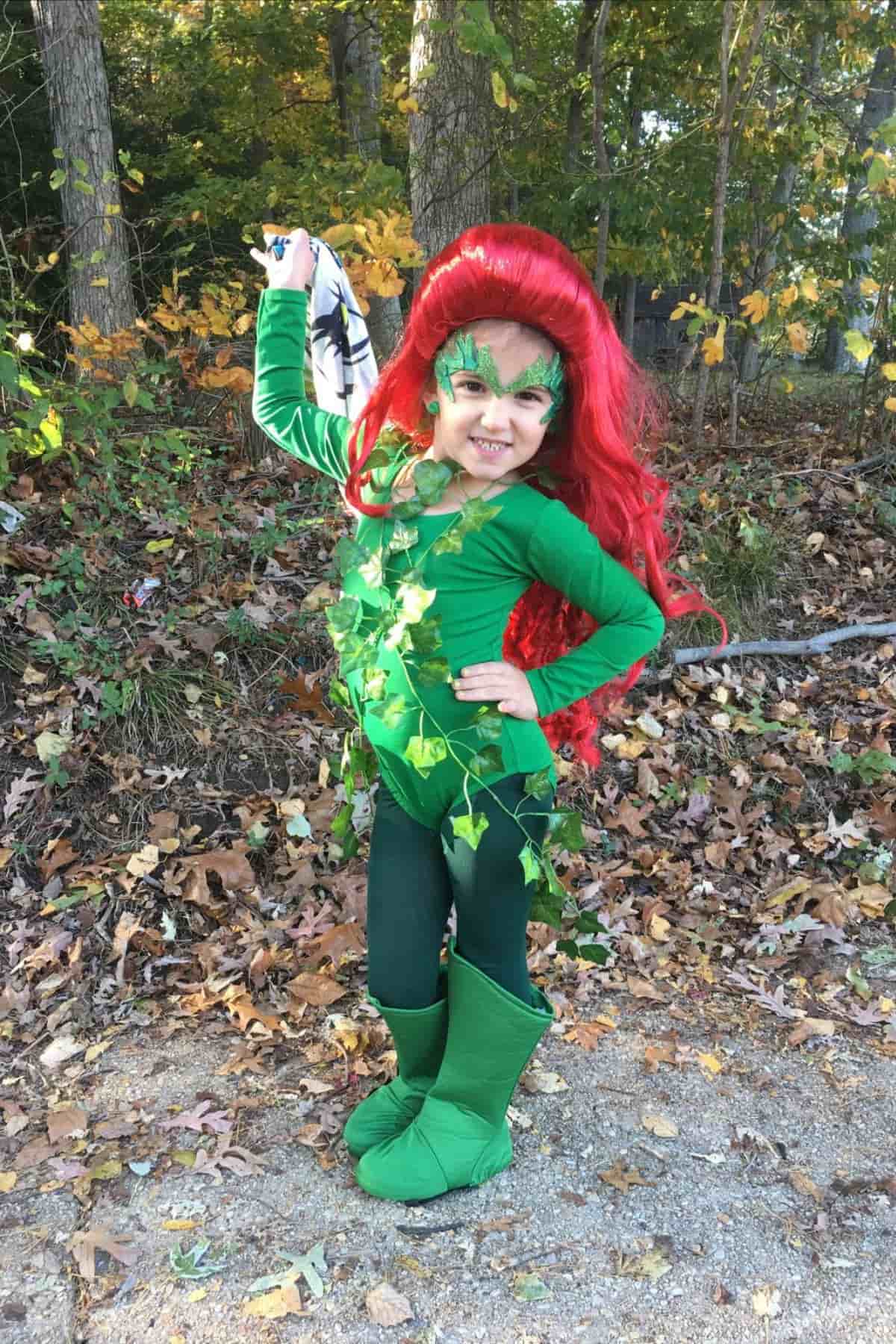 Child in a green costume with red wig and leafy decorations poses outdoors, holding a cloth with black design, surrounded by autumn leaves and trees.