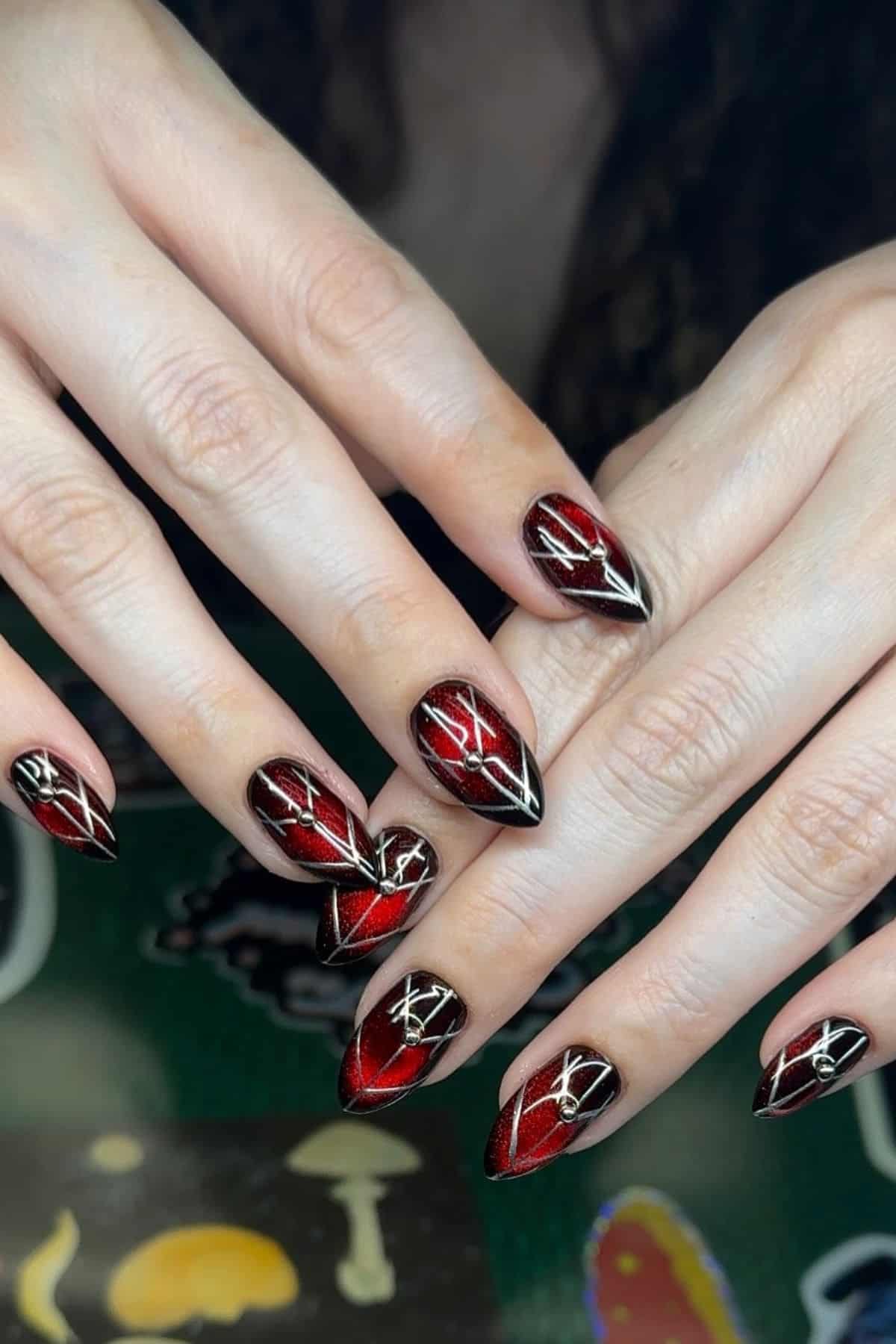 Close-up of hands with long, pointed nails painted in black and red ombre, featuring intricate white geometric line designs on each nail.