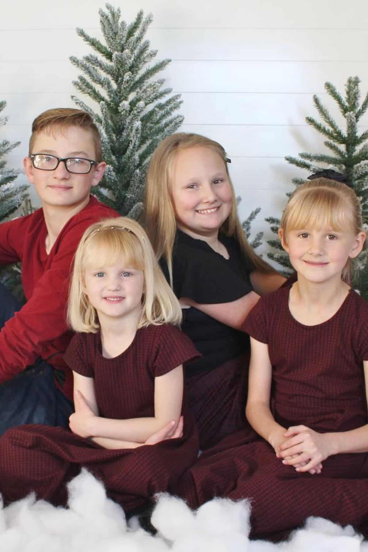 Four children sit in front of artificial snow and pine trees, with three girls in matching maroon dresses and a boy in a maroon shirt and glasses, all smiling at the camera.