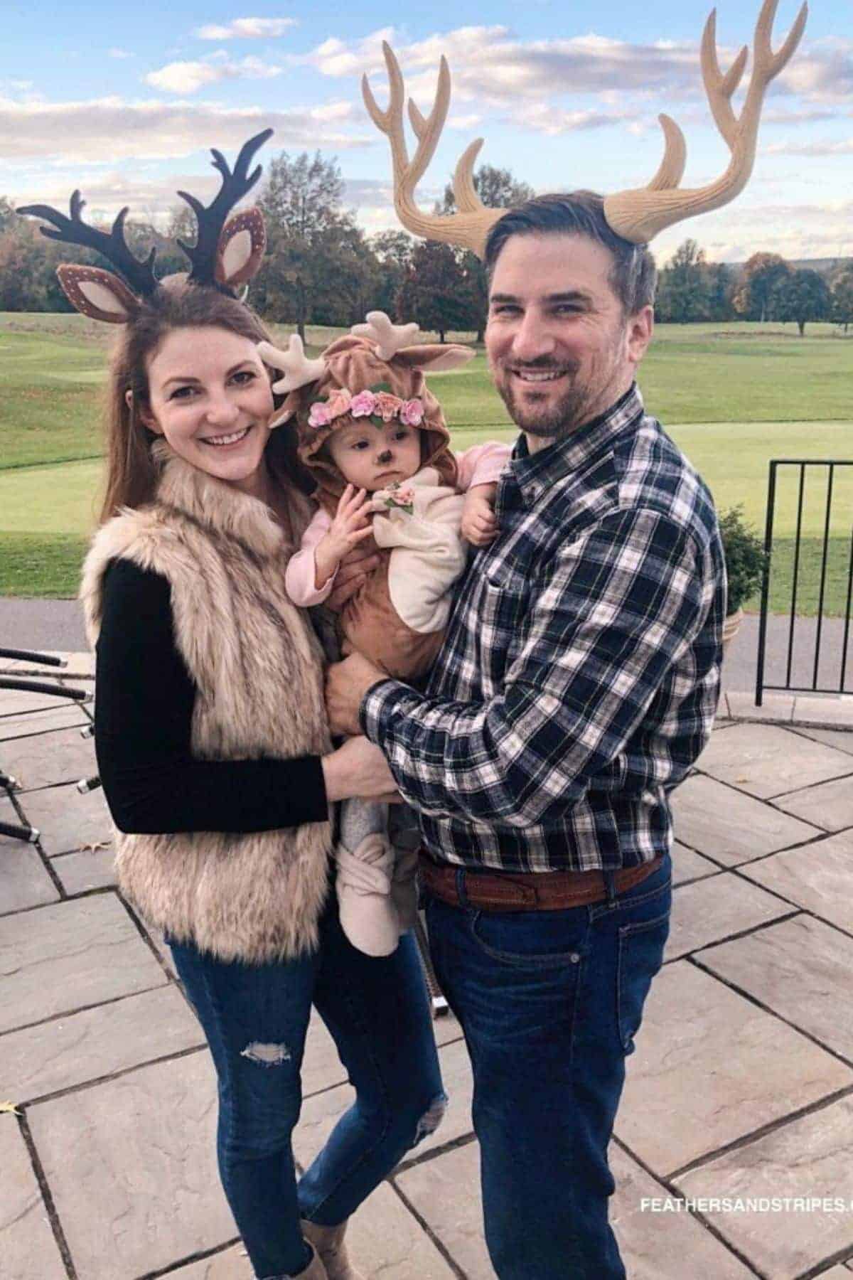 Three people stand outside wearing antler headbands; two adults hold a baby dressed in a deer-themed costume. Trees and a grassy field are in the background.
