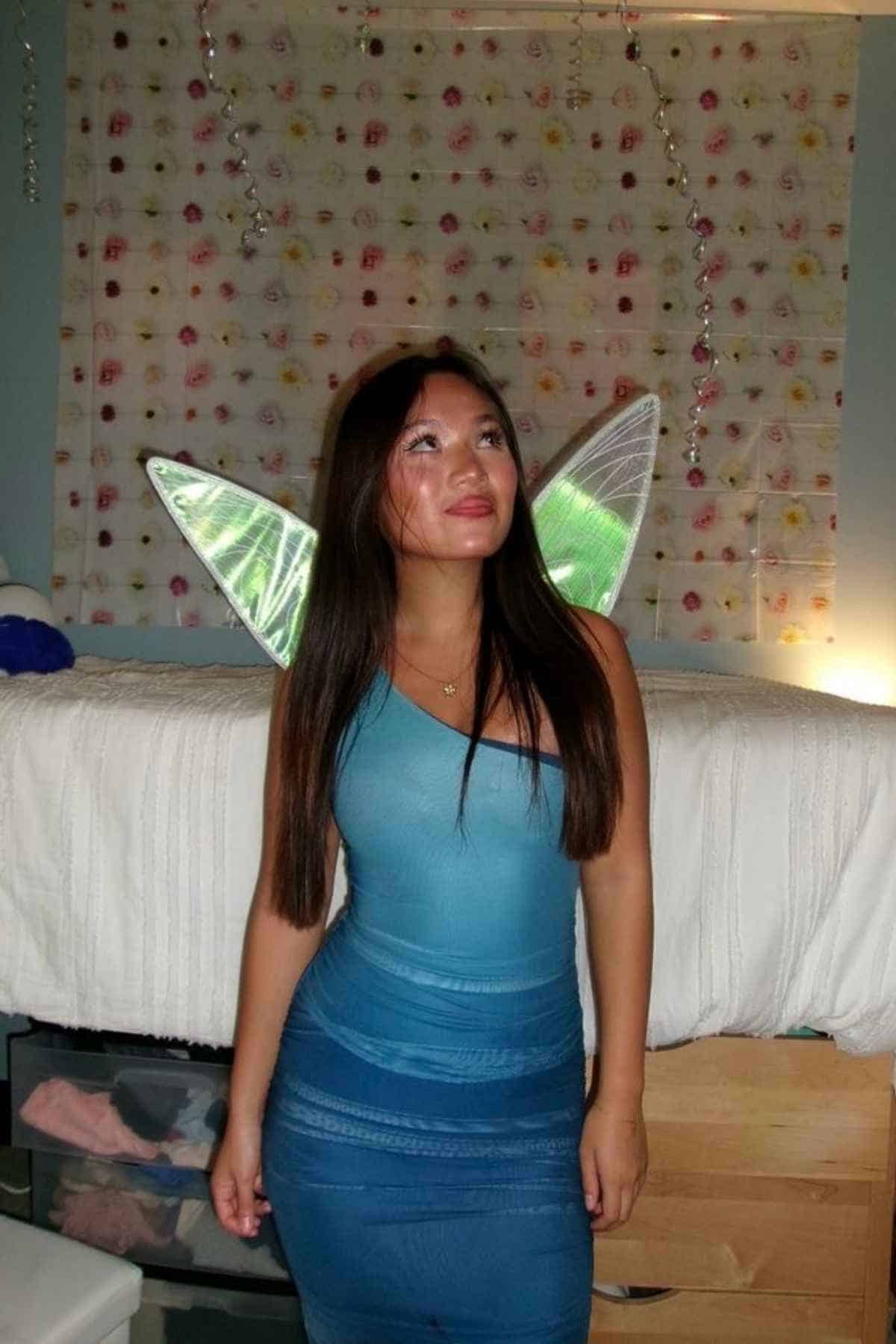 Woman in a blue dress stands indoors wearing shiny fairy wings, in front of a bed with a flower-patterned wall hanging in the background.