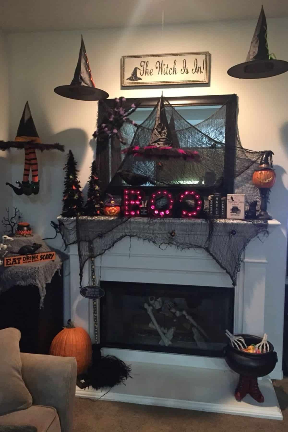 A fireplace decorated for Halloween with witch hats, spider webs, pumpkins, skeletons, and a “BOO” sign; a “The Witch Is In” sign hangs above the mantel.