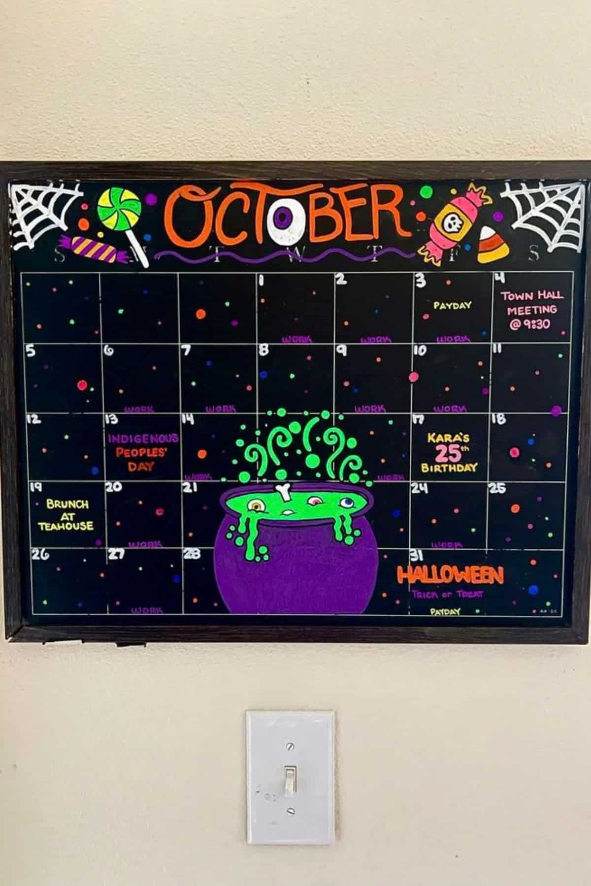 Black dry-erase calendar for October with Halloween decorations. Events such as payday, meetings, and holidays are written in neon colors. A large bubbling cauldron is drawn in the center.
