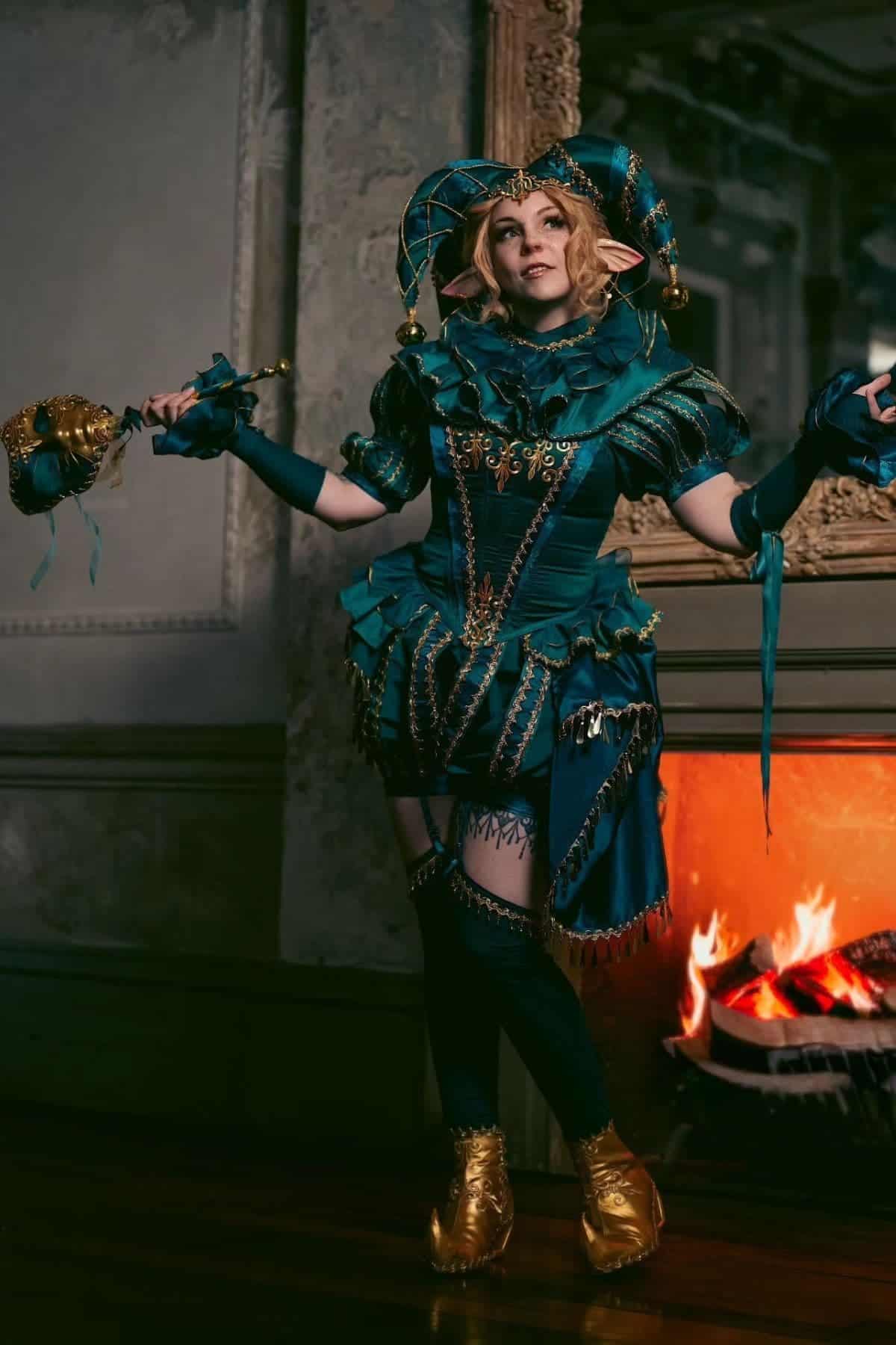 Person in elaborate teal and gold jester costume with pointed hat, elf ears, and holding a decorative staff, standing indoors by a lit fireplace.