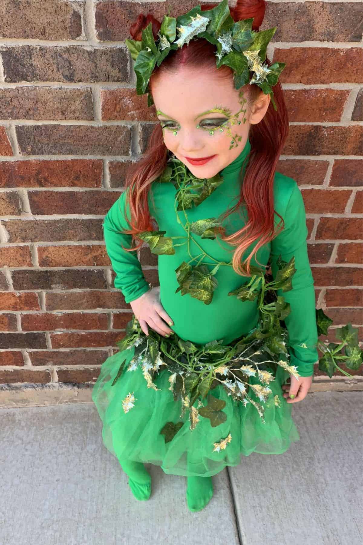 Young child in a green costume with leaf decorations stands by a brick wall, wearing green tights, a leafy crown, and green face paint.