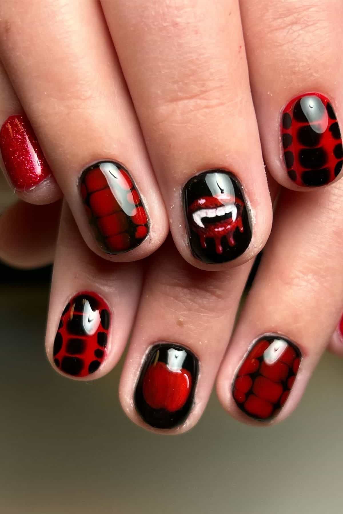 Fingernails painted with black and red designs, including spider webs, an apple, and vampire fangs with blood on the middle nail.