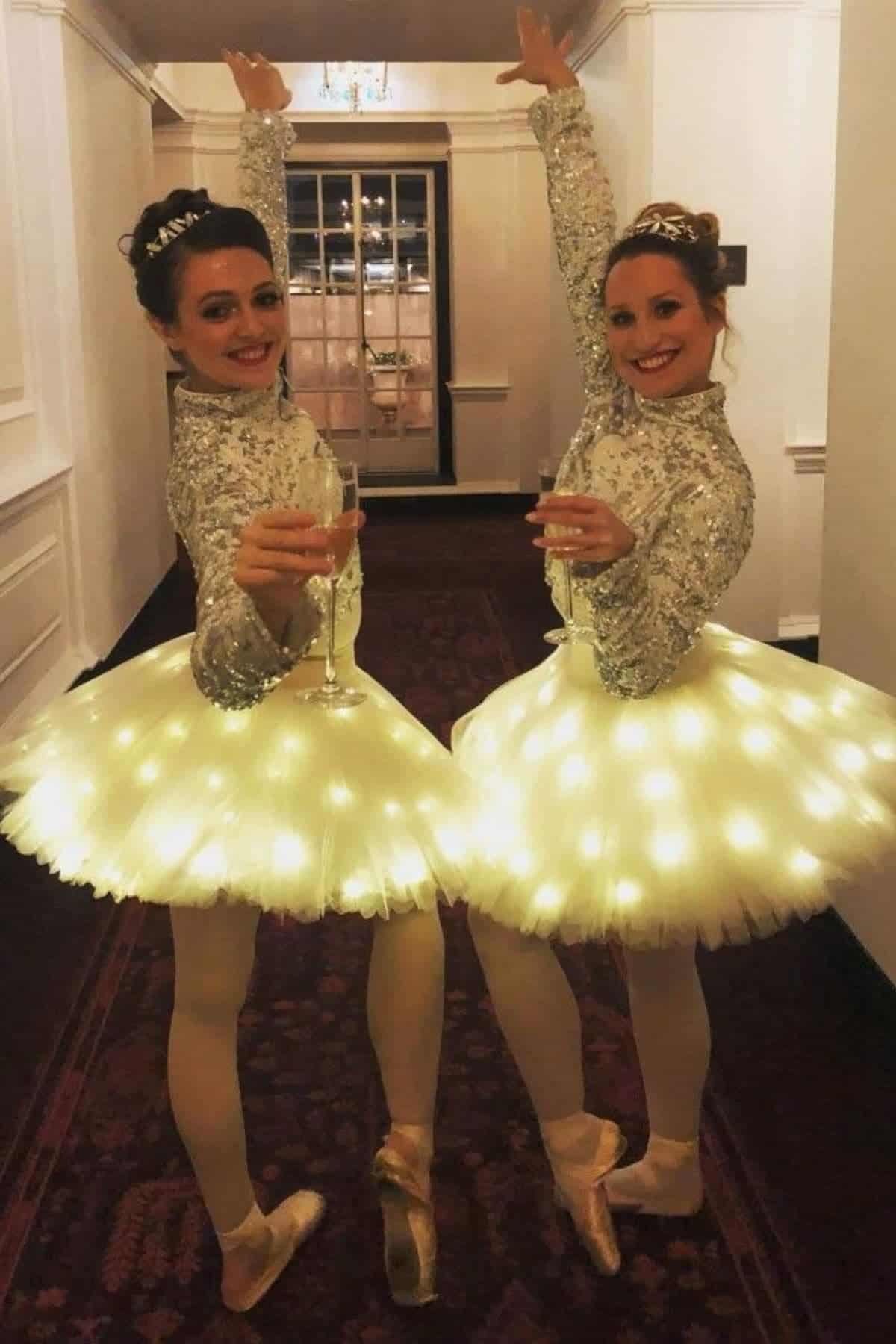Two women in sparkling ballet costumes with illuminated tutus stand on pointe and hold champagne glasses, smiling at the camera in a hallway.