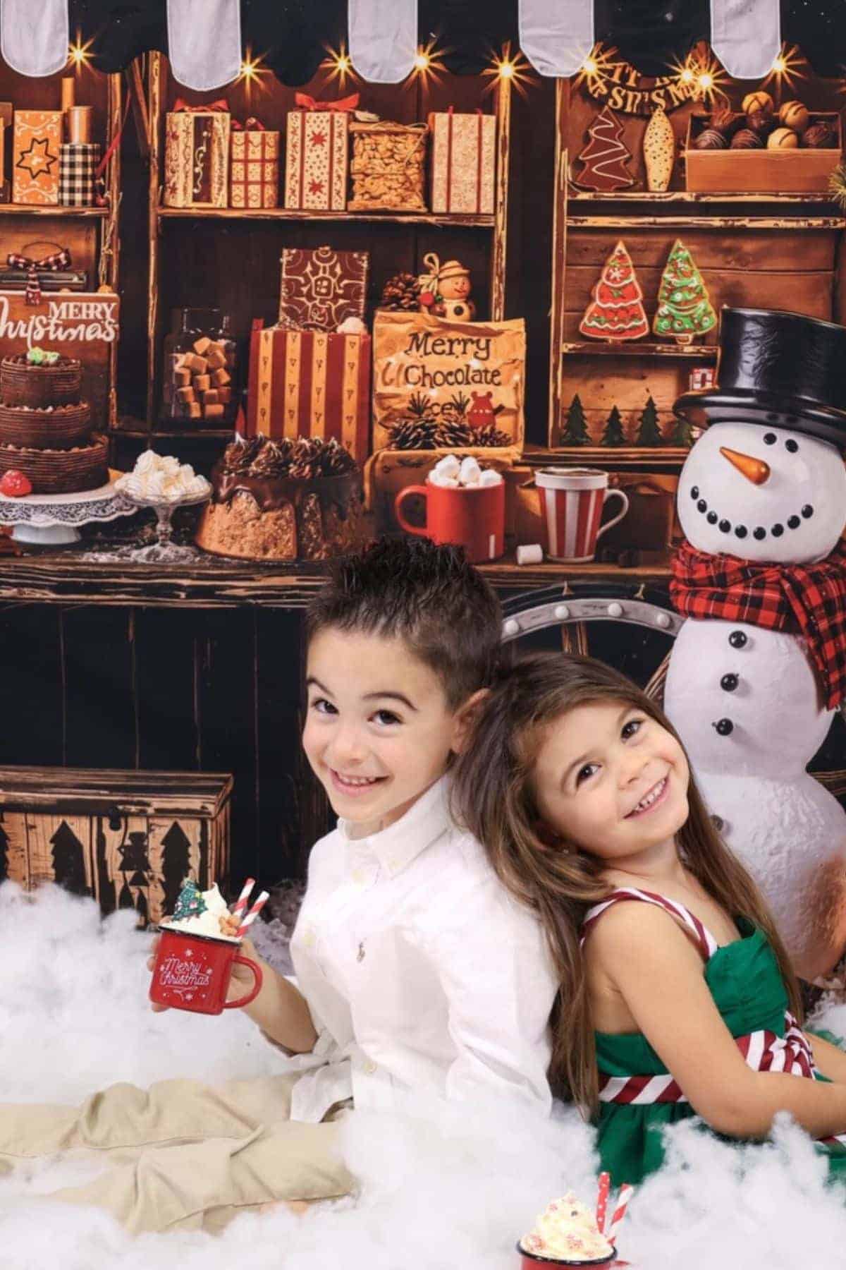 Two children sit on fake snow, smiling and holding holiday mugs in front of a festive Christmas-themed backdrop with desserts, decorations, and a snowman.