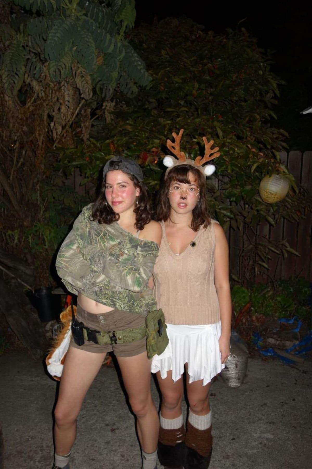 Two women stand outside at night; one wears camouflage with a utility belt, and the other is dressed as a deer with antlers and face paint. Both pose for the camera.