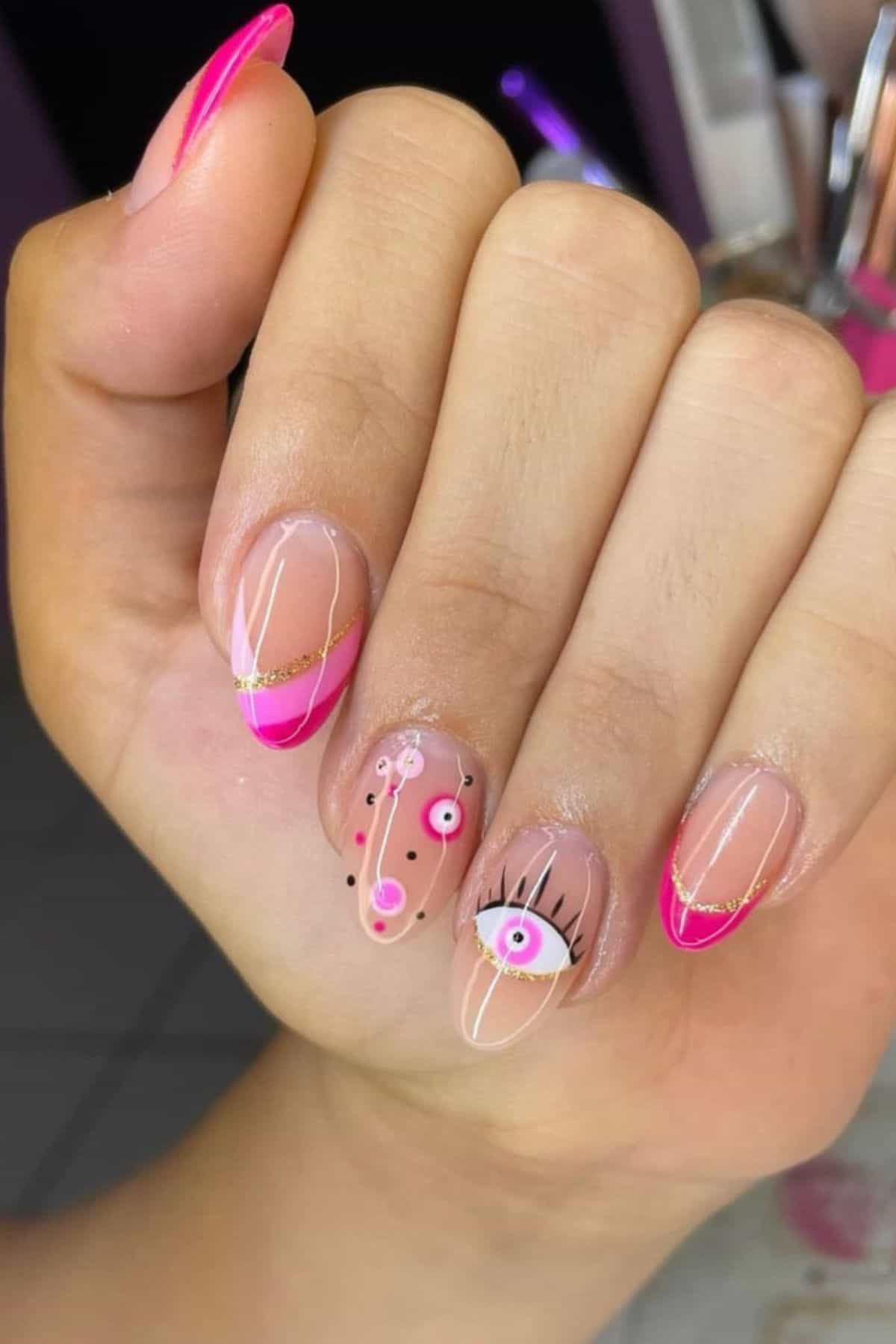 A hand with almond-shaped nails featuring pink French tips, gold lines, and abstract eye and face designs on a nude base.