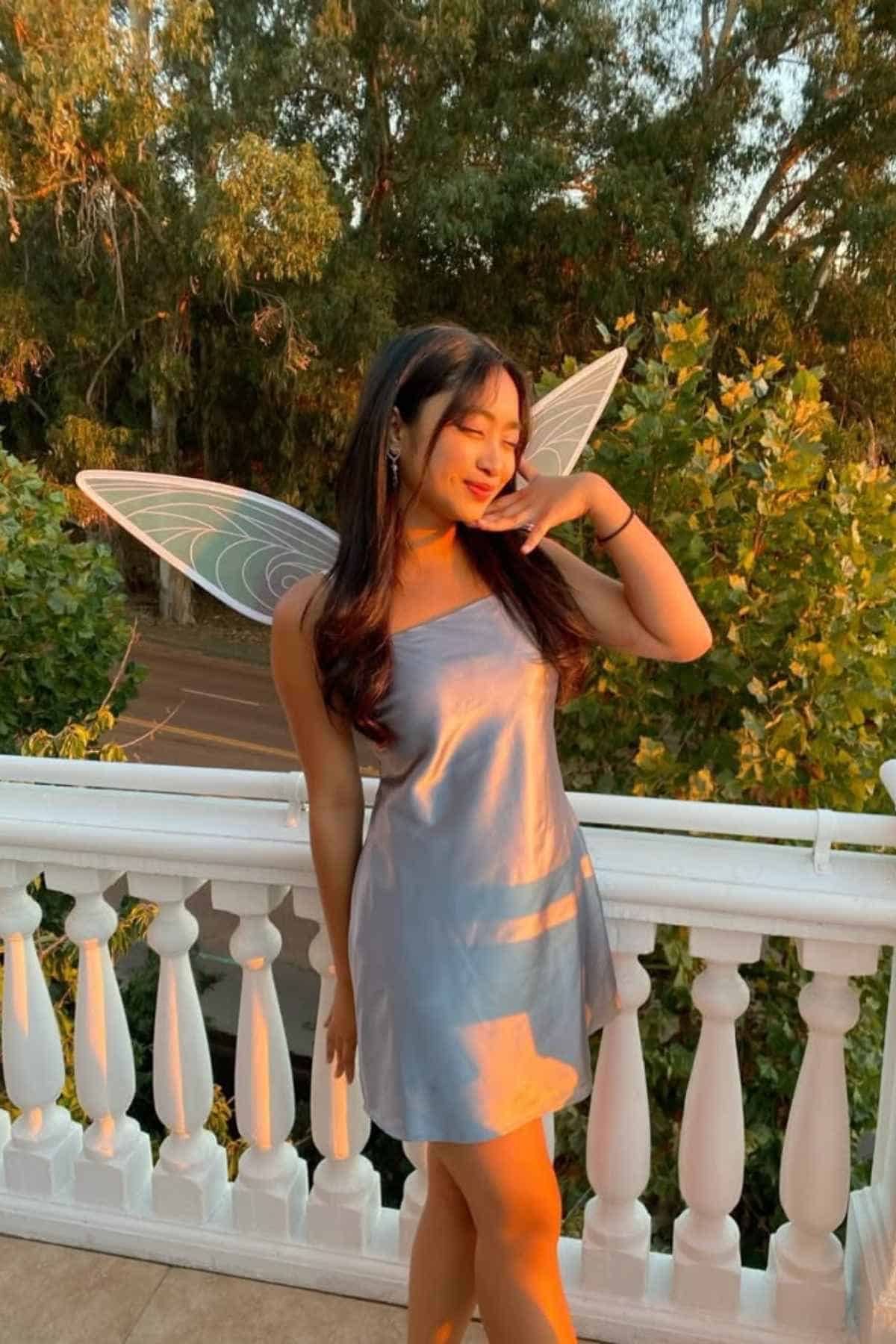 A woman in a light blue dress and fairy wings stands on a balcony with white railings, surrounded by green trees and evening sunlight.
