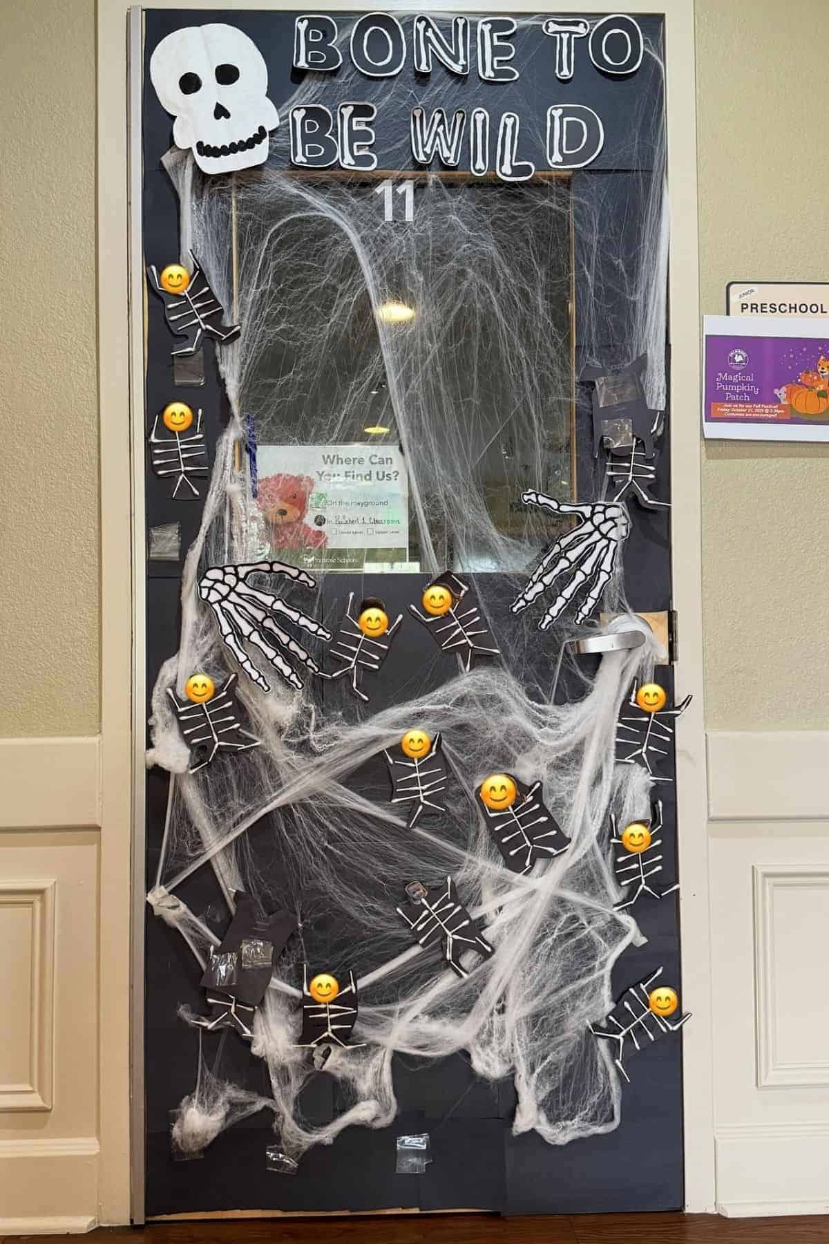Classroom door decorated with a “Bone to Be Wild” skeleton theme, featuring fake cobwebs, skeleton cutouts with emoji faces, and a large paper skull at the top.
