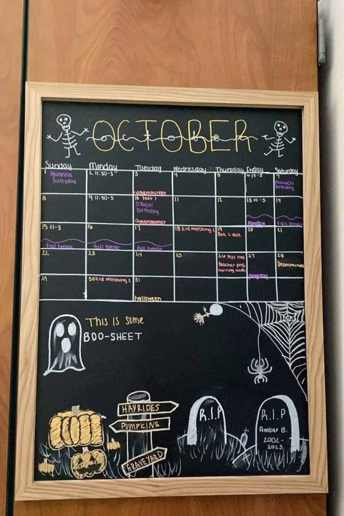 A blackboard calendar for October is decorated with Halloween drawings, including skeletons, pumpkins, gravestones, a spider web, and a ghost saying “This is some boo-sheet.”.