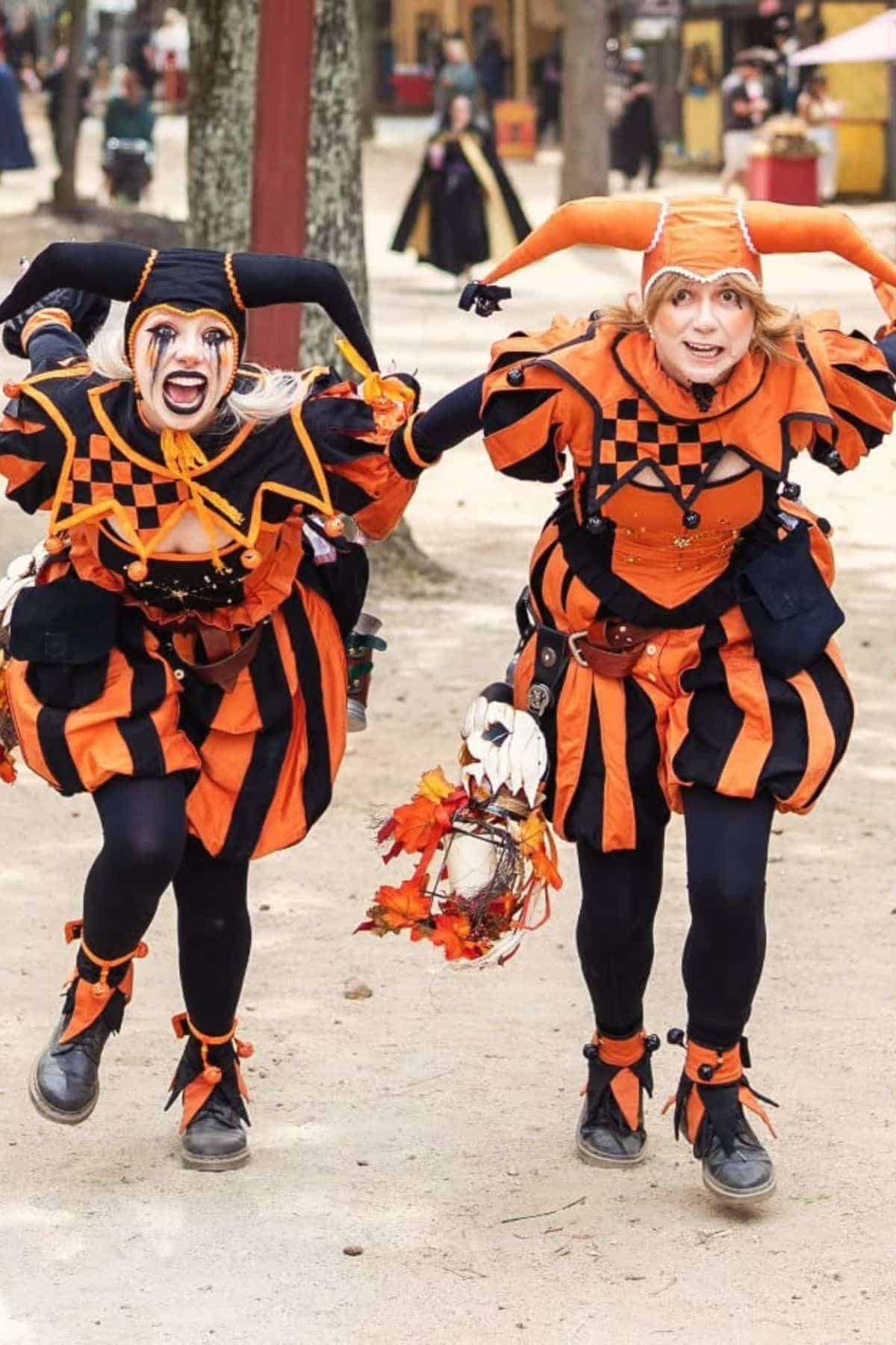 Two people dressed as jesters in orange and black costumes walk together outdoors, holding hands and smiling, with one carrying a decorative lantern.