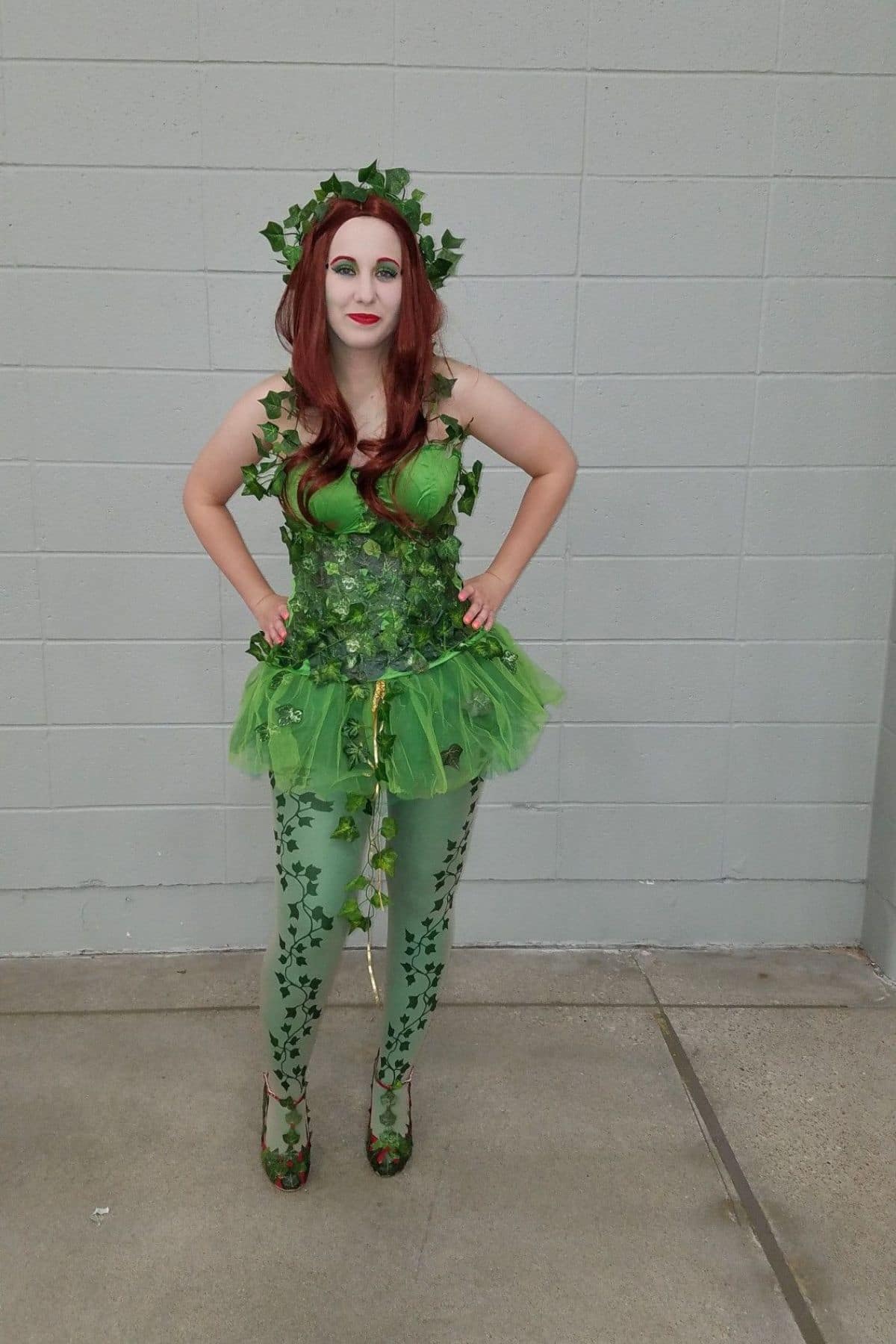 Person in a green leafy costume with red hair, green tights, and green shoes stands against a gray cinder block wall.