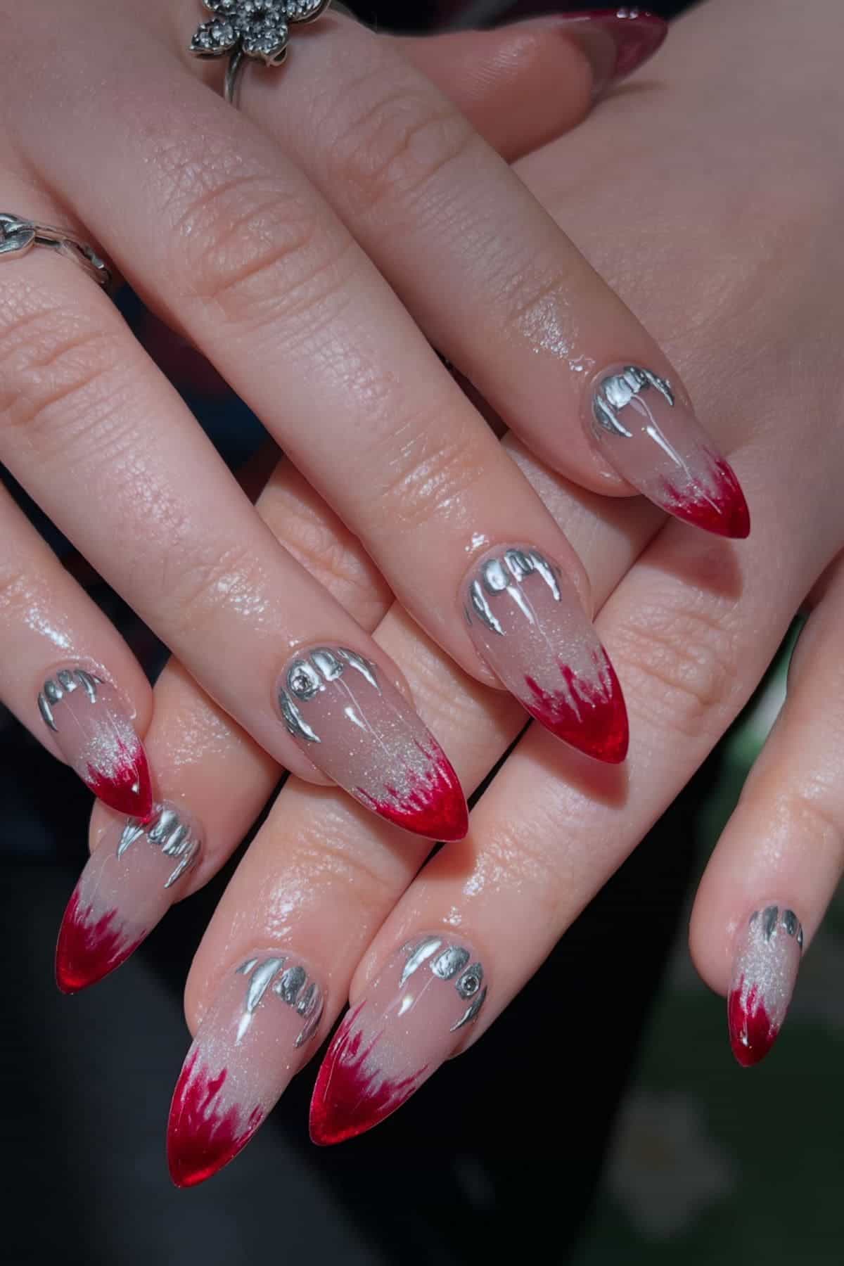 Hands with long, pointed nails featuring silver drip designs near the cuticles and red gradient tips resembling blood.