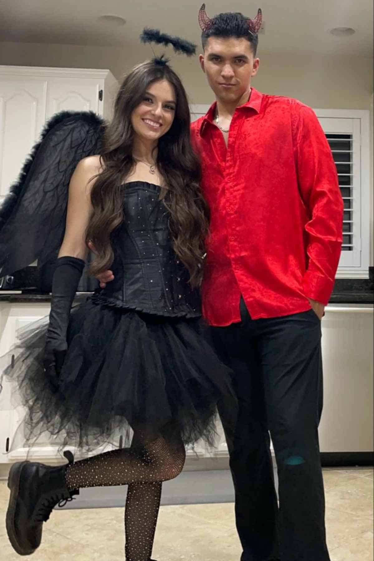 Two people in a kitchen; one dressed as a dark angel with black wings, and the other as a devil in a red shirt with small red horns.
