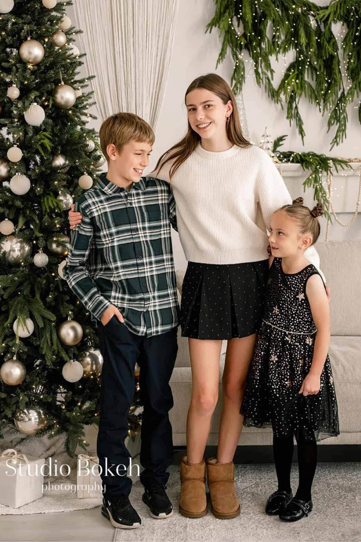 Three children stand next to a decorated Christmas tree and a sofa, smiling for a holiday photo in a living room.